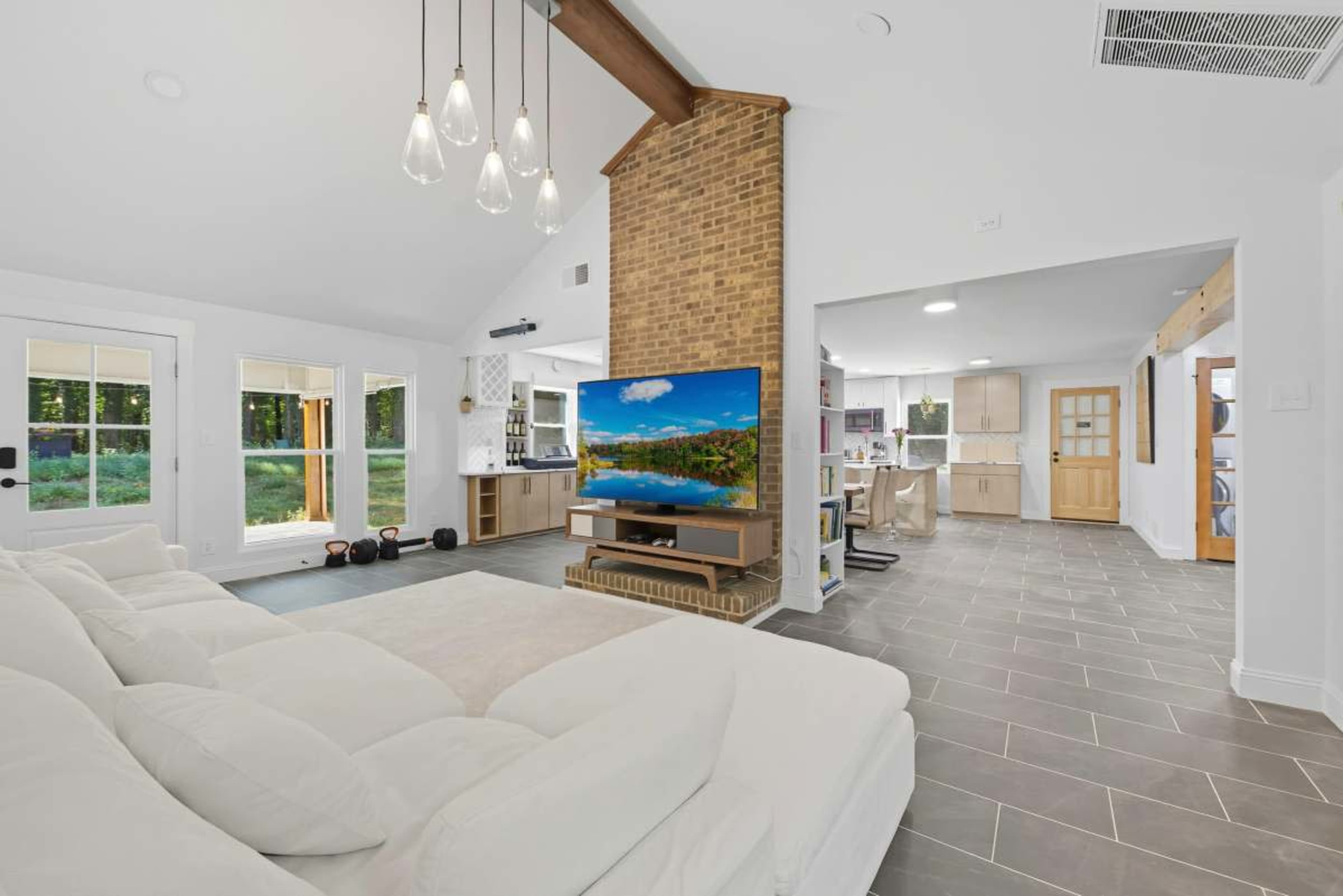 The image shows a modern living area featuring a large white sofa, a brick fireplace, and a flat-screen TV, with a view into a bright kitchen area.