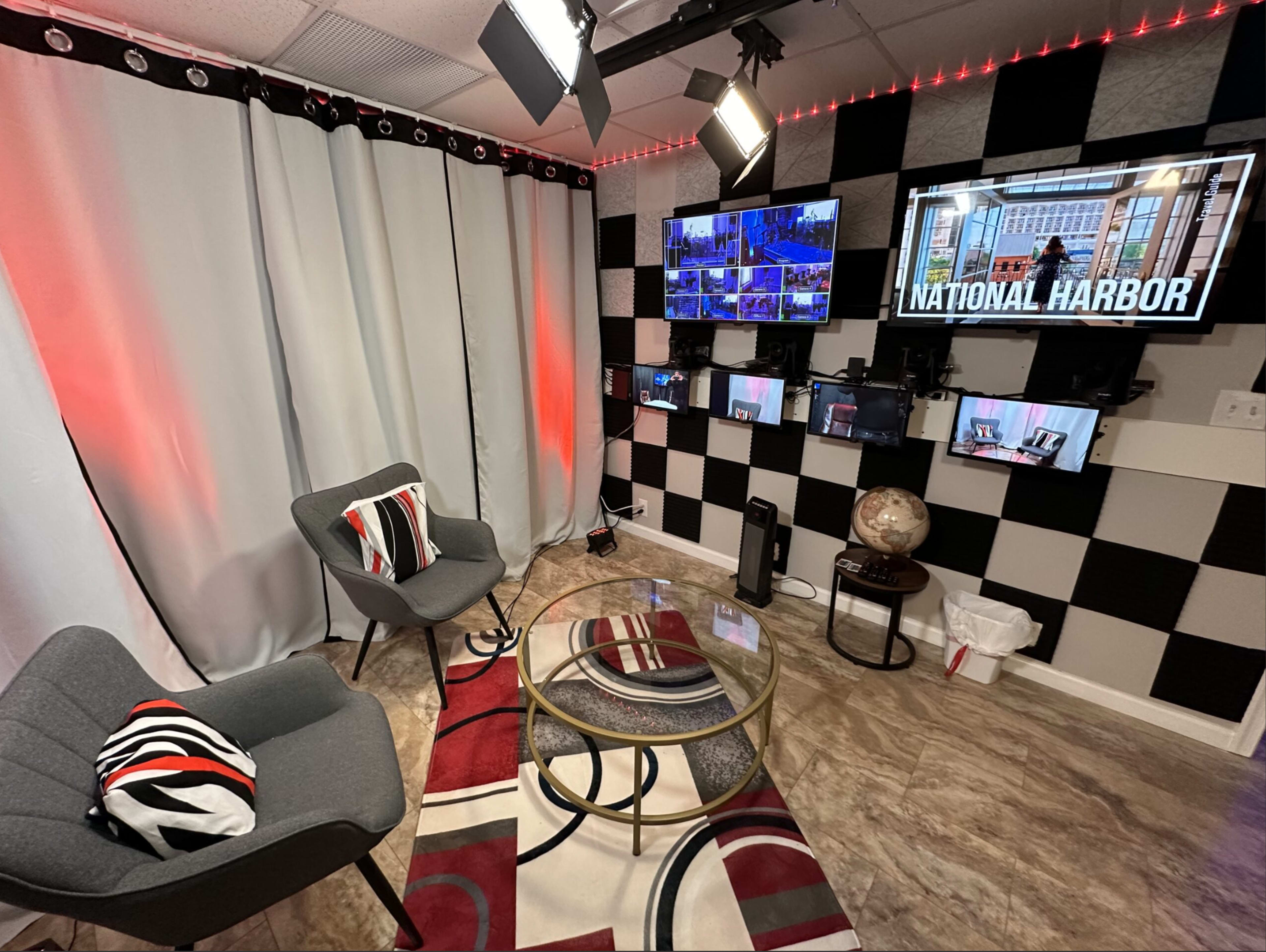 A modern studio setup with two chairs, a round glass table, multiple screens displaying a location, and a large wall covered in black and white checkered tiles.