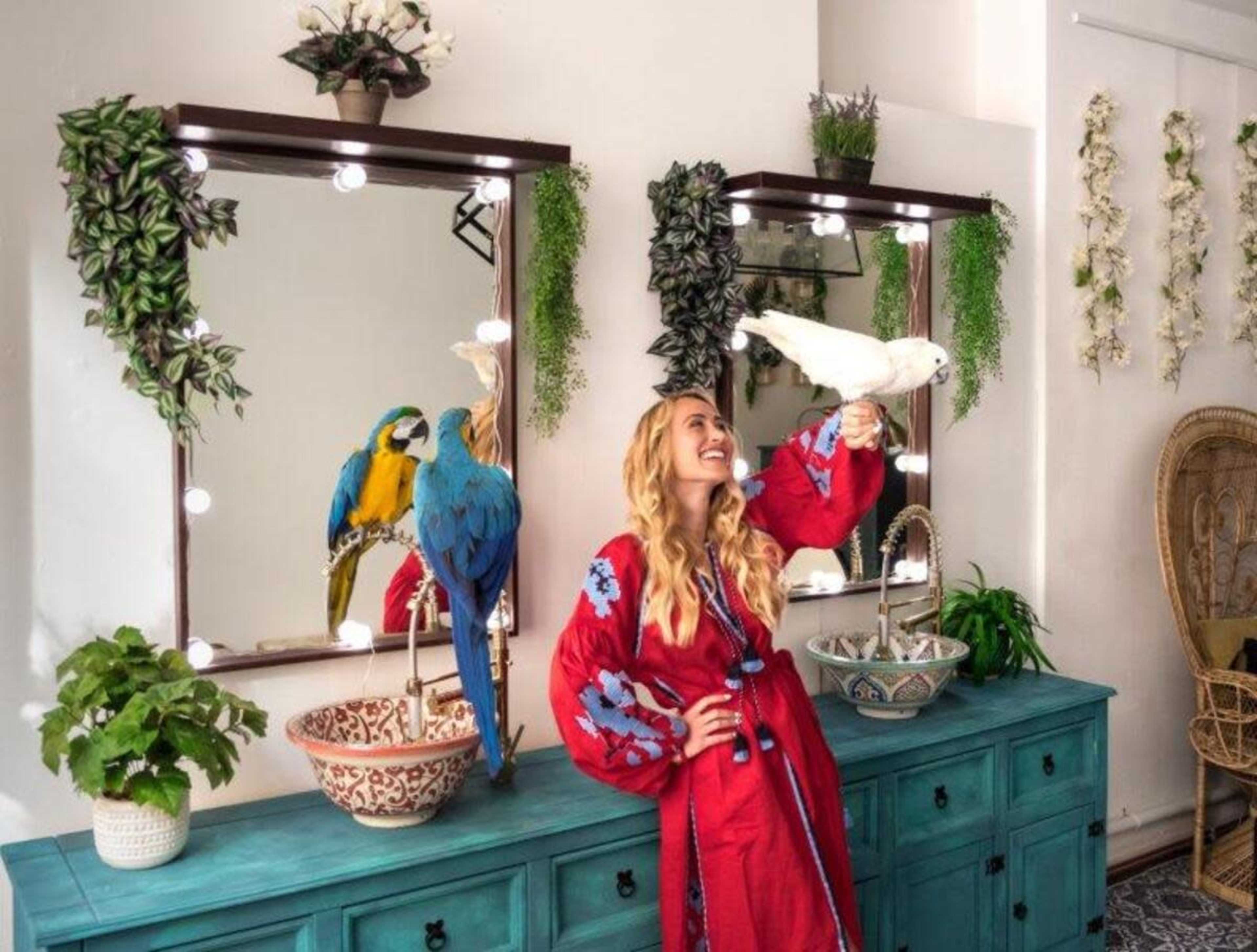 A woman in a red robe stands in front of a mirror, holding a white bird, while colorful parrots perch on a nearby cabinet.