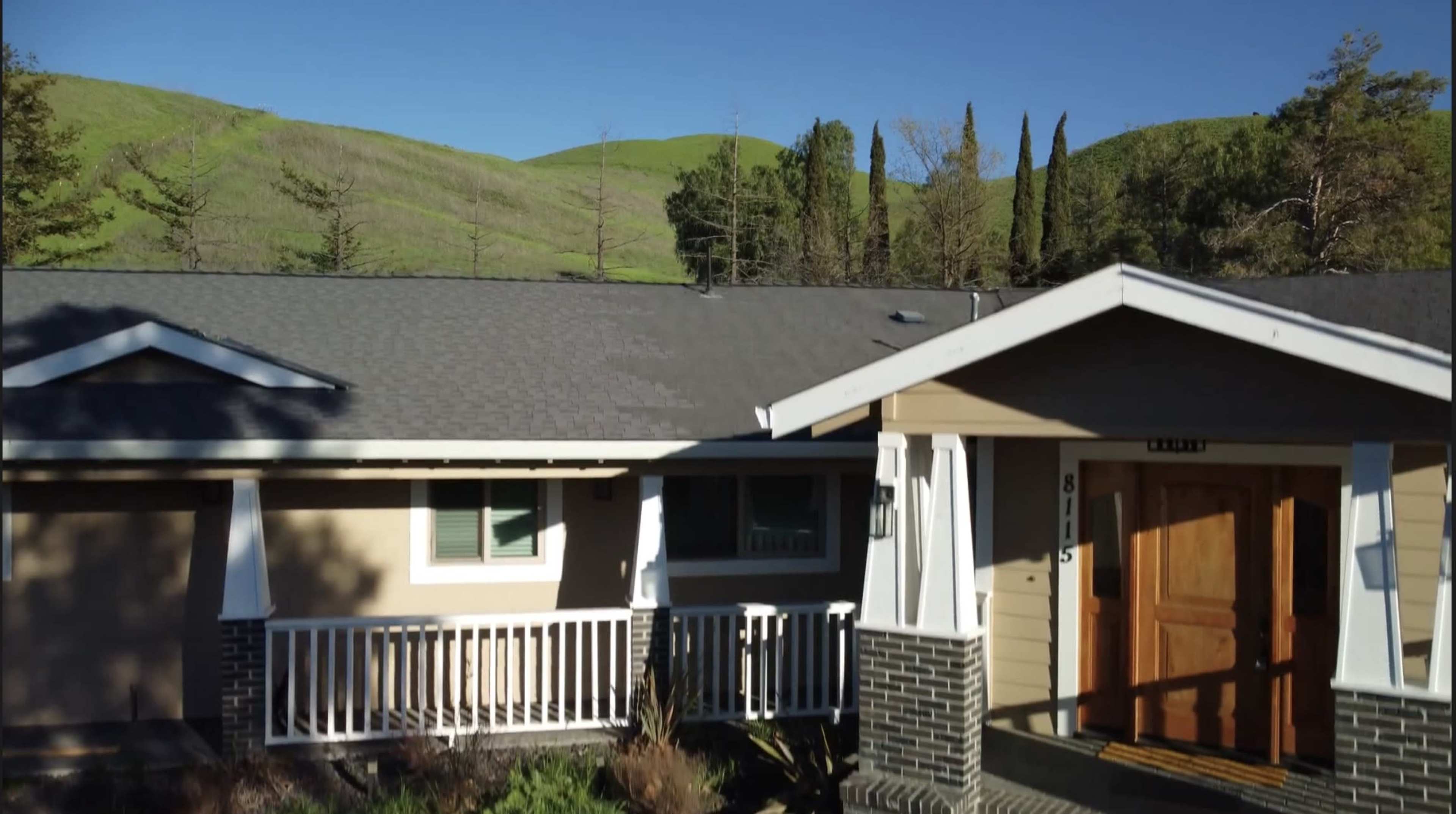 The image shows a single-story house with a porch, surrounded by green hills and trees.