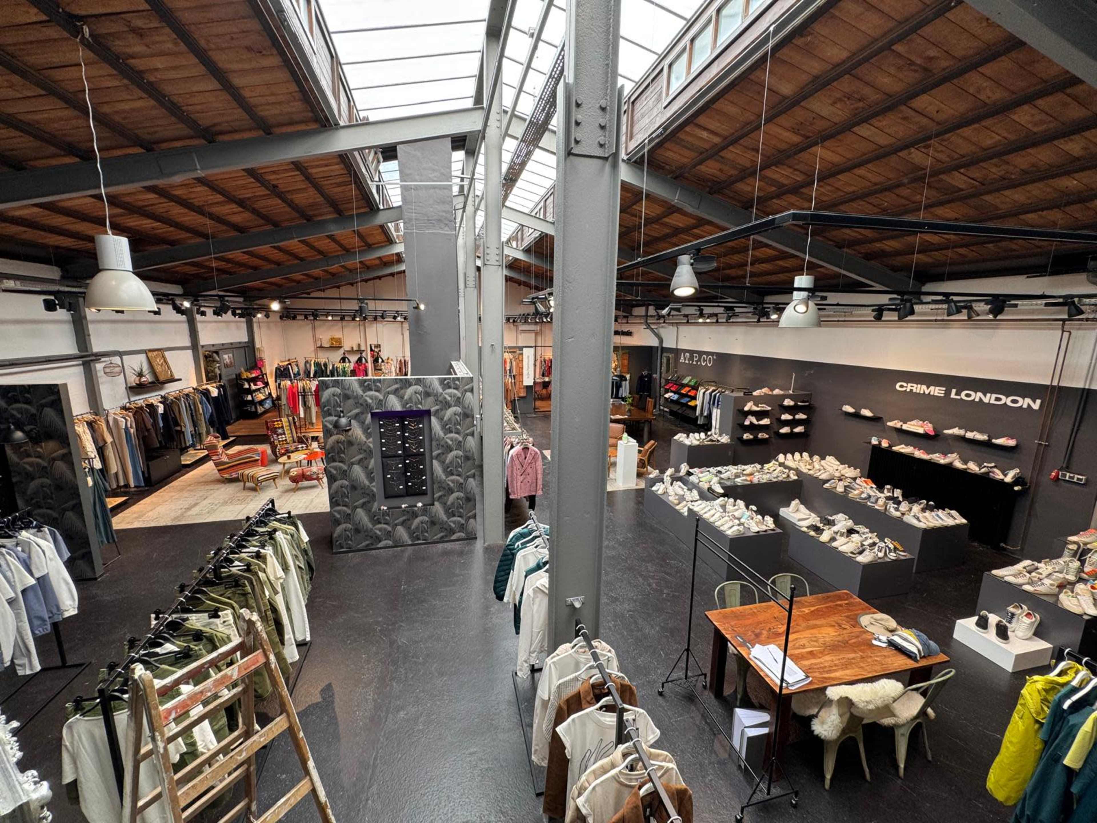 The image shows an expansive clothing store with a variety of garments displayed on racks and shelves, featuring a central seating area and a well-lit ceiling with large windows.