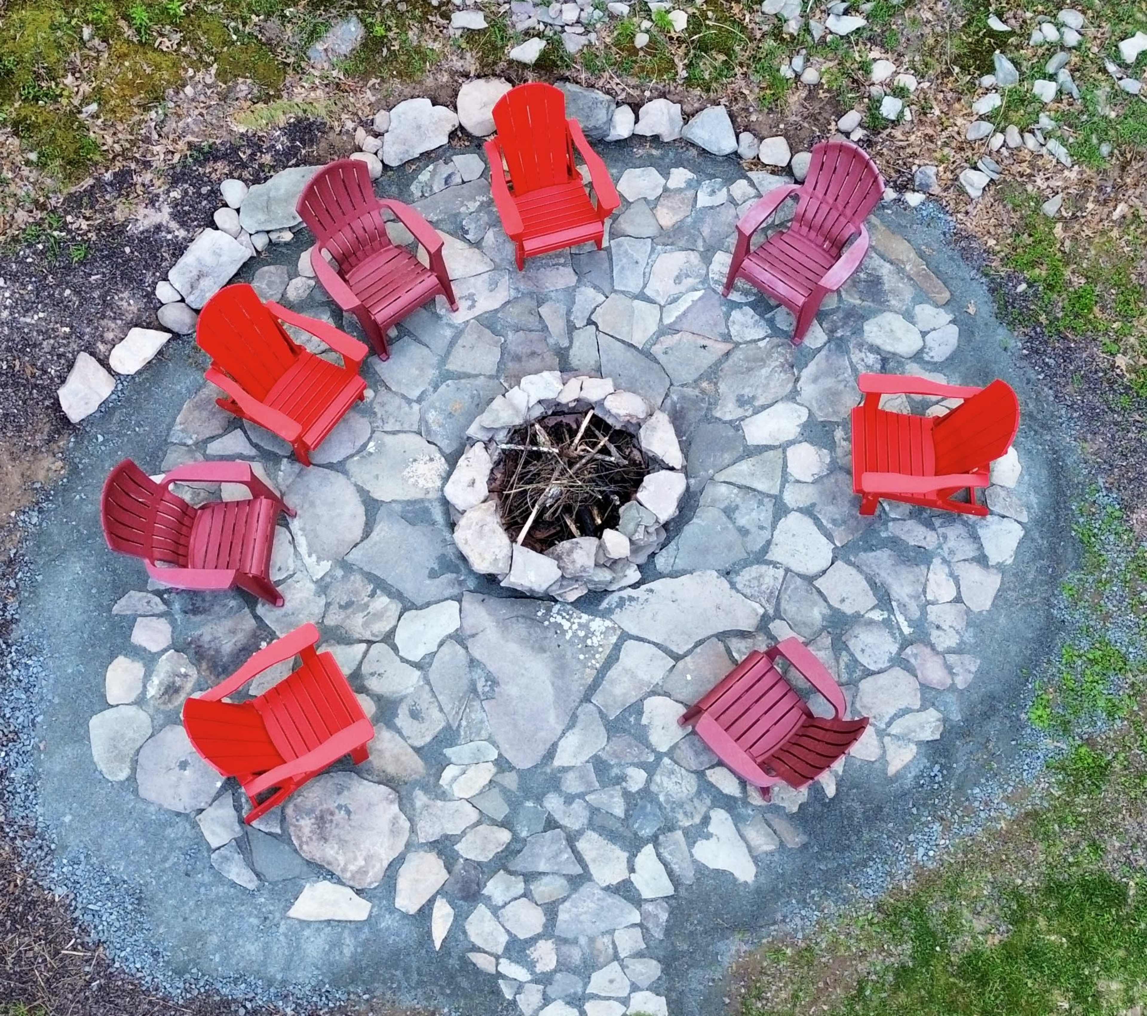 A circular arrangement of six red adirondack chairs surrounds a stone fire pit in a grassy area.