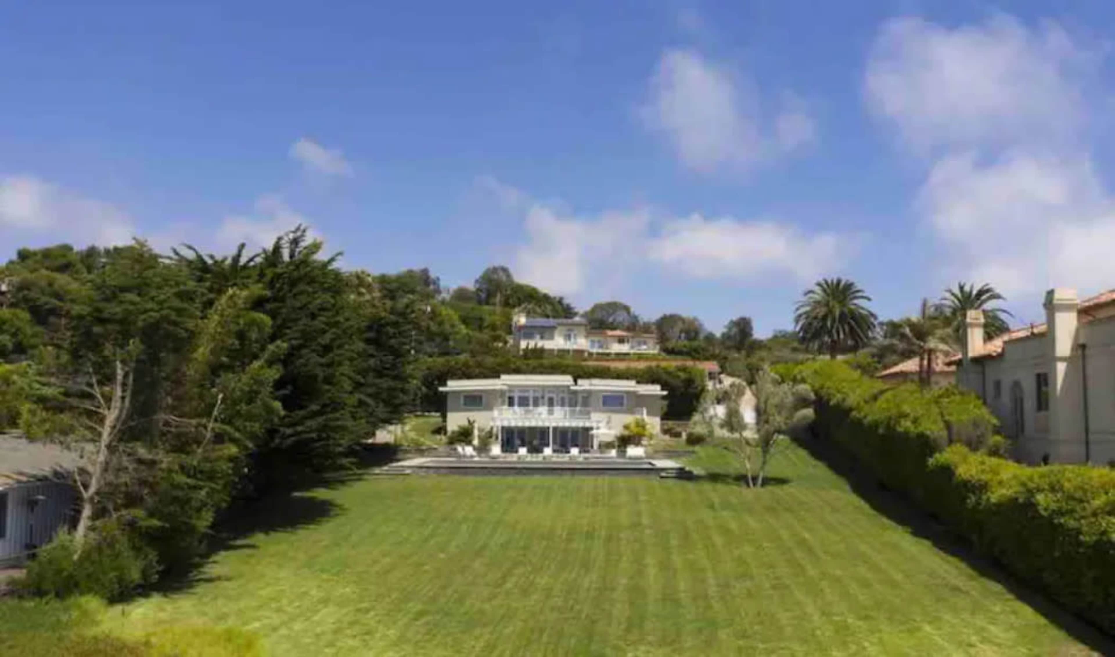 A modern house sits at the center of a large green lawn, surrounded by trees and other homes on a hillside under a clear blue sky.