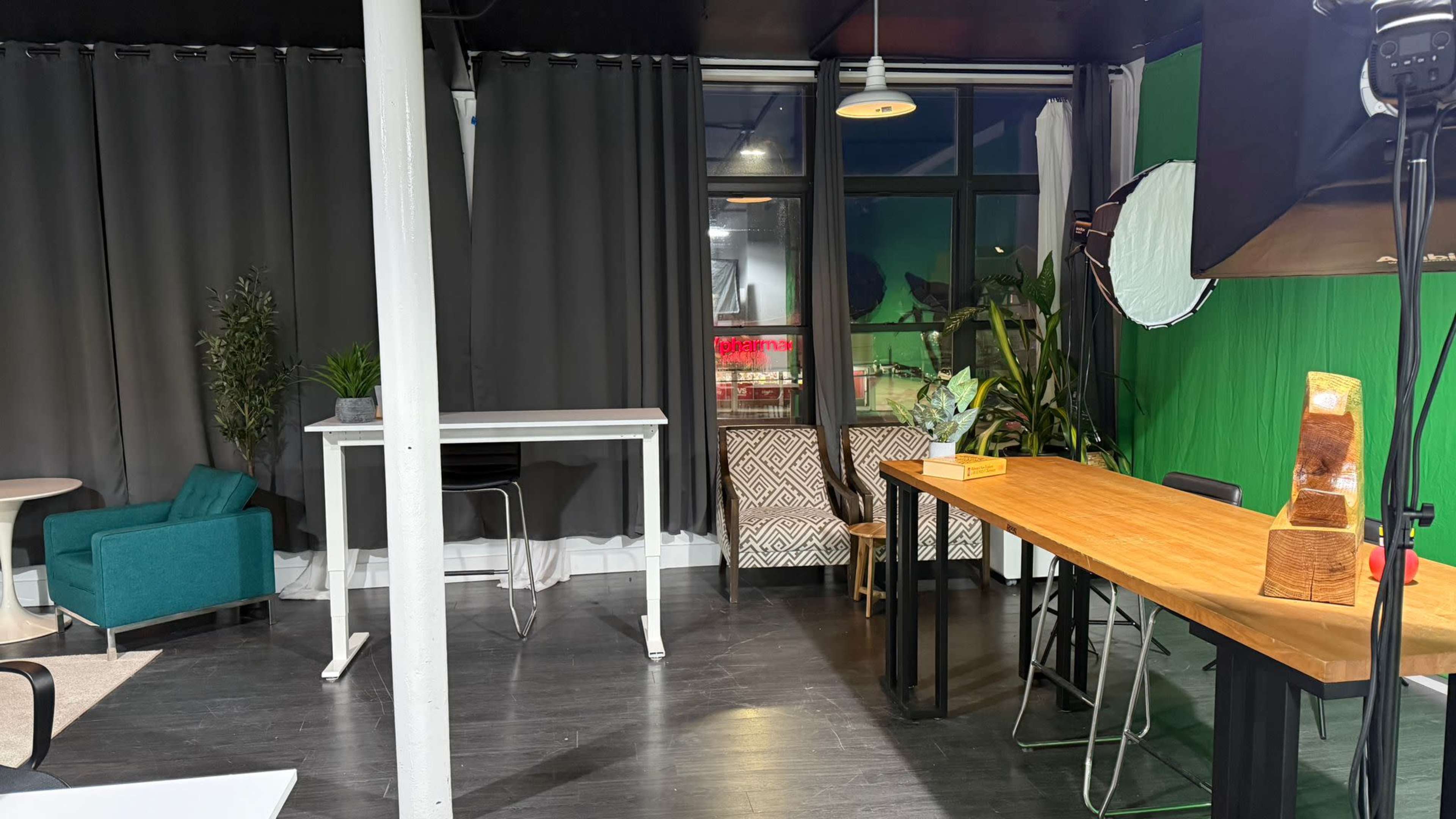 The image shows a modern studio space with a wooden table, two chairs, and a green screen backdrop, alongside plants and a partial view of a large window.