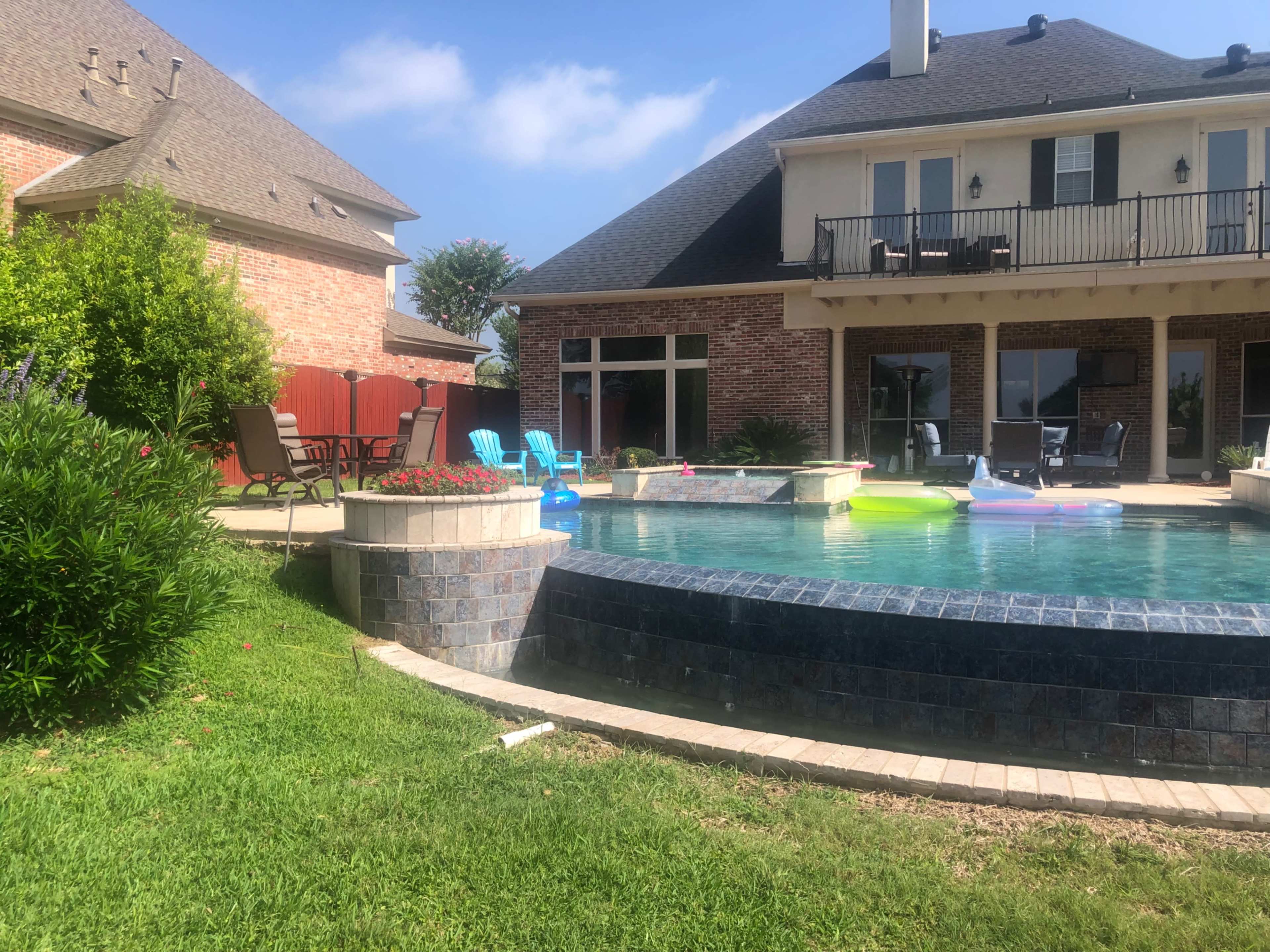 A backyard pool area with a brick house, lounge chairs, and colorful inflatable floaties in the water.