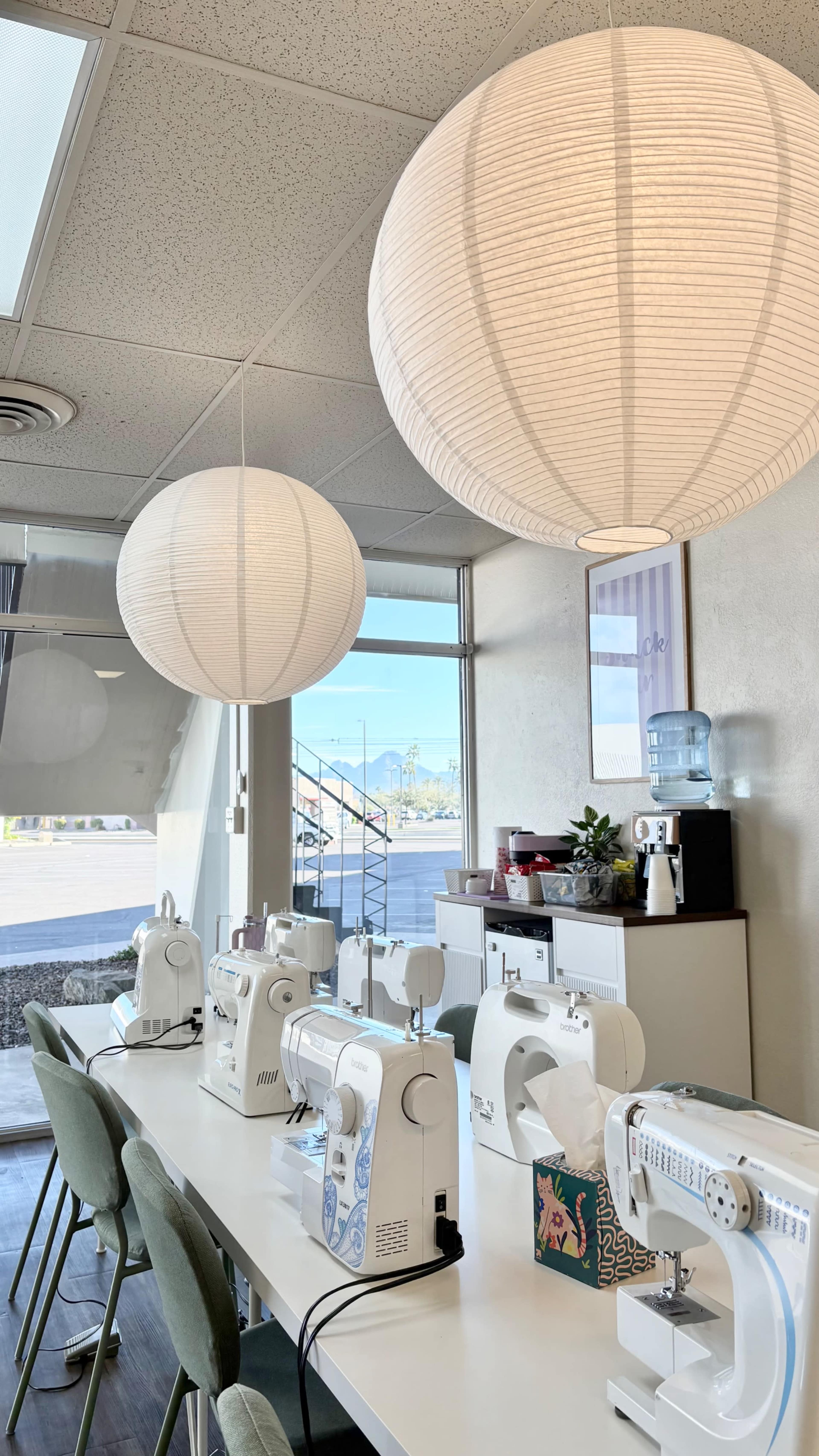 The image shows a bright sewing studio with multiple sewing machines arranged on a table, illuminated by large paper lanterns hanging from the ceiling.