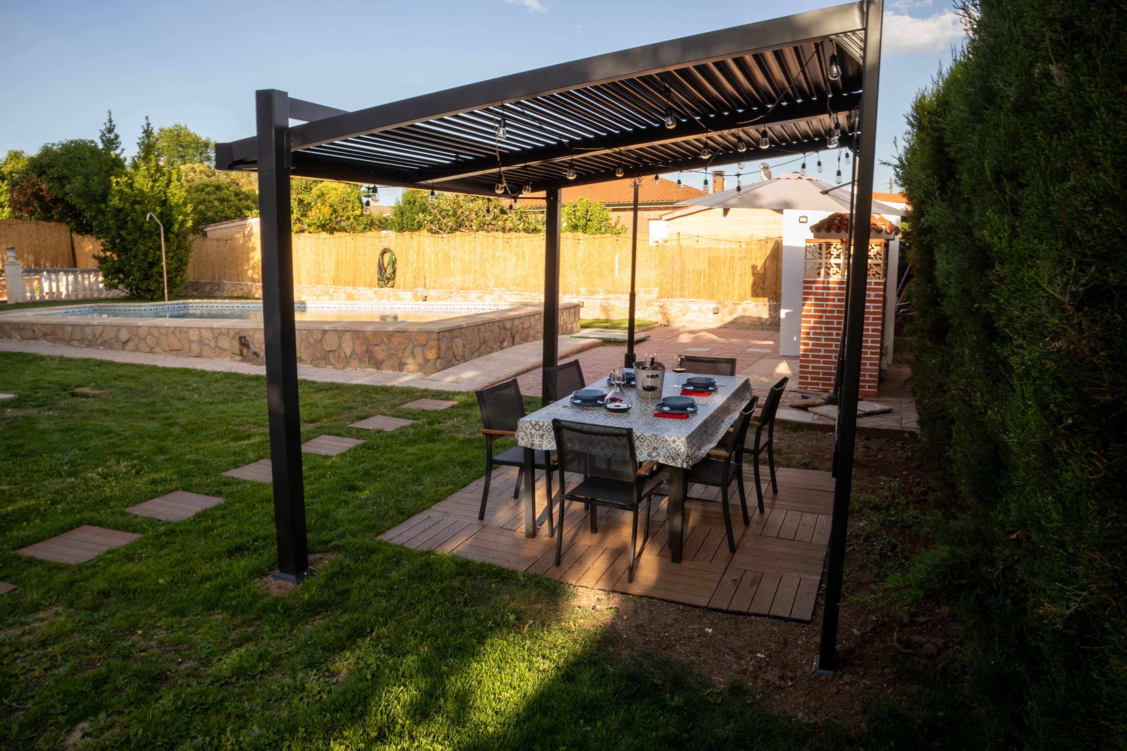 A dining table with chairs is set under a pergola in a backyard, adjacent to a stone pool.