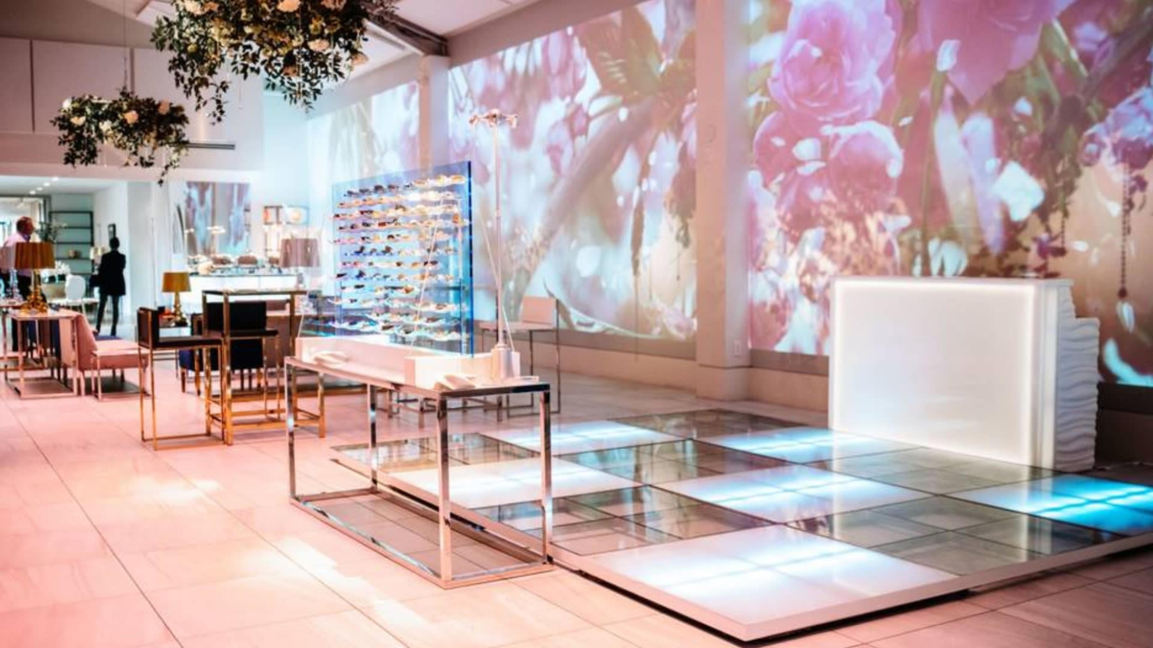 The image depicts a modern event space featuring a sleek bar, glass display stands, and projected floral visuals on the walls.