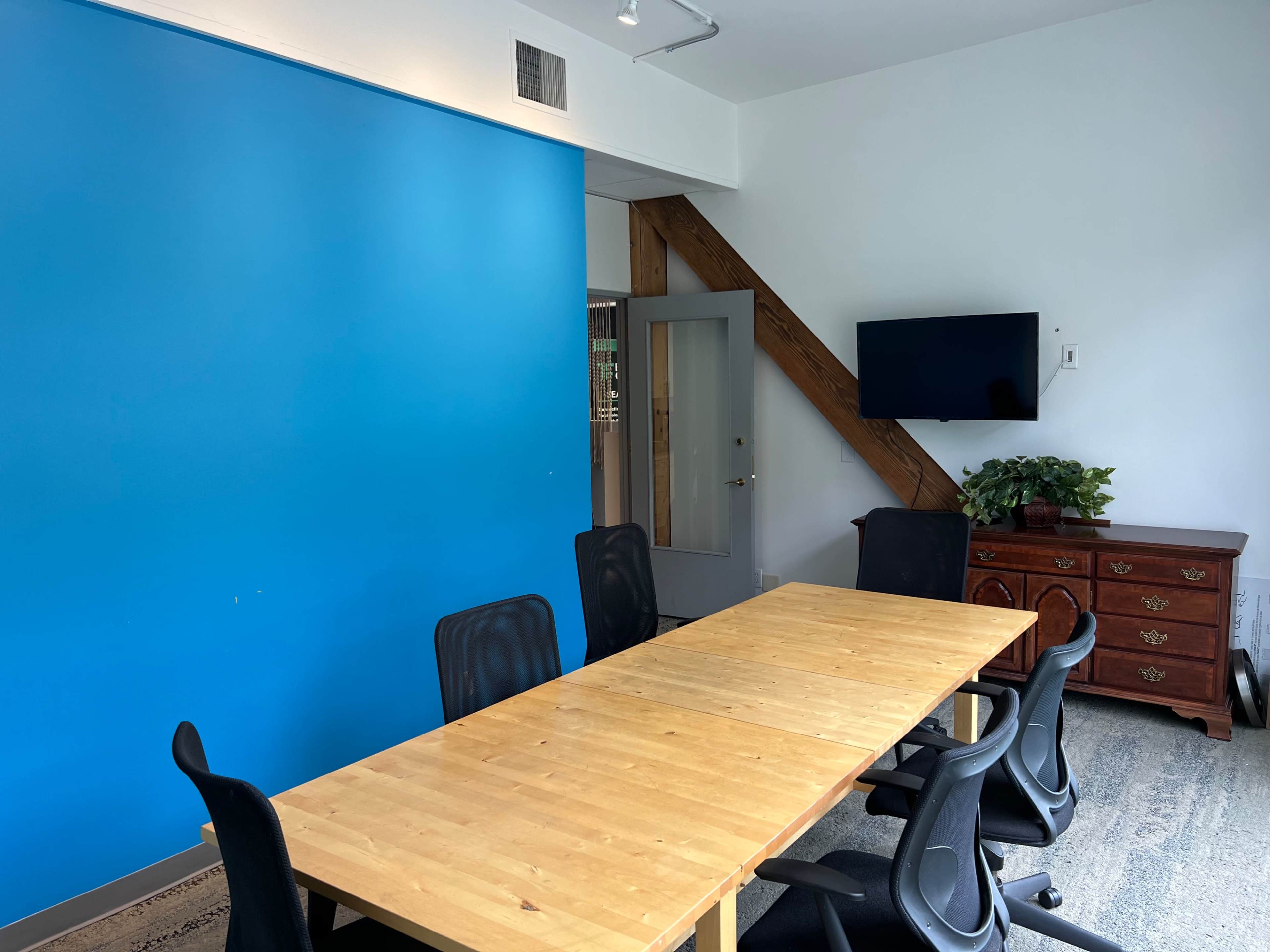A conference room features a large wooden table surrounded by black chairs, with a blue wall and a television mounted nearby.