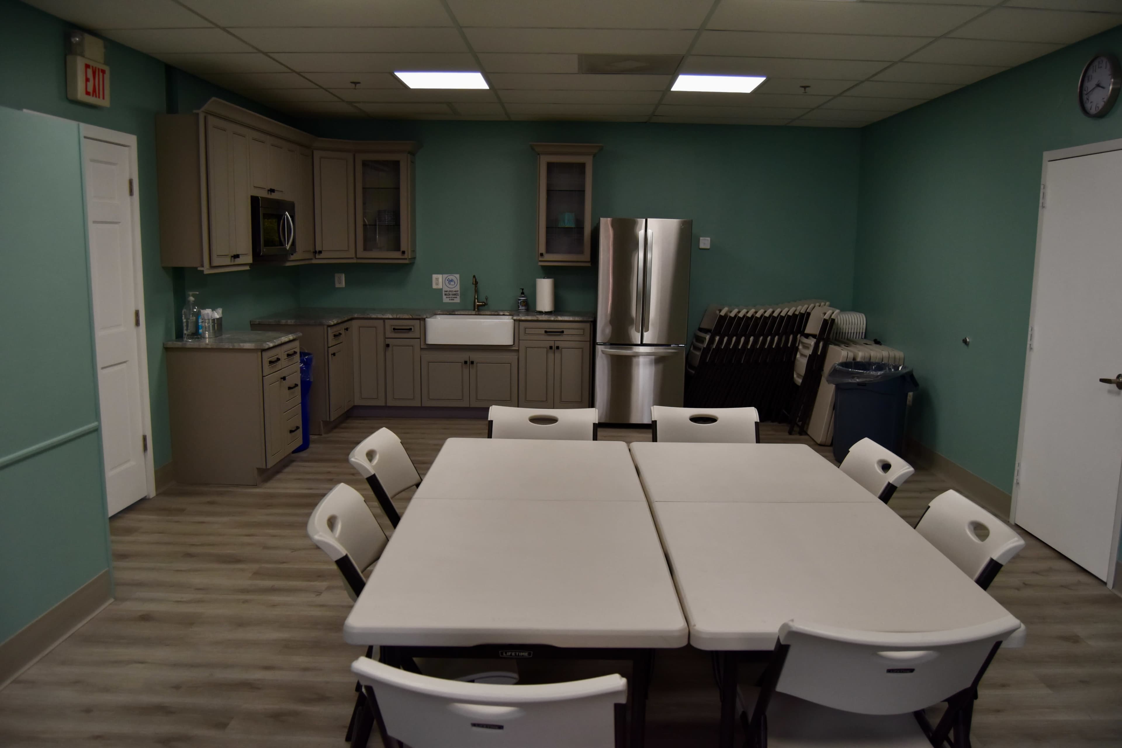 A kitchenette area with light green walls, a stainless steel refrigerator, and a set of foldable tables and chairs arranged in the center.