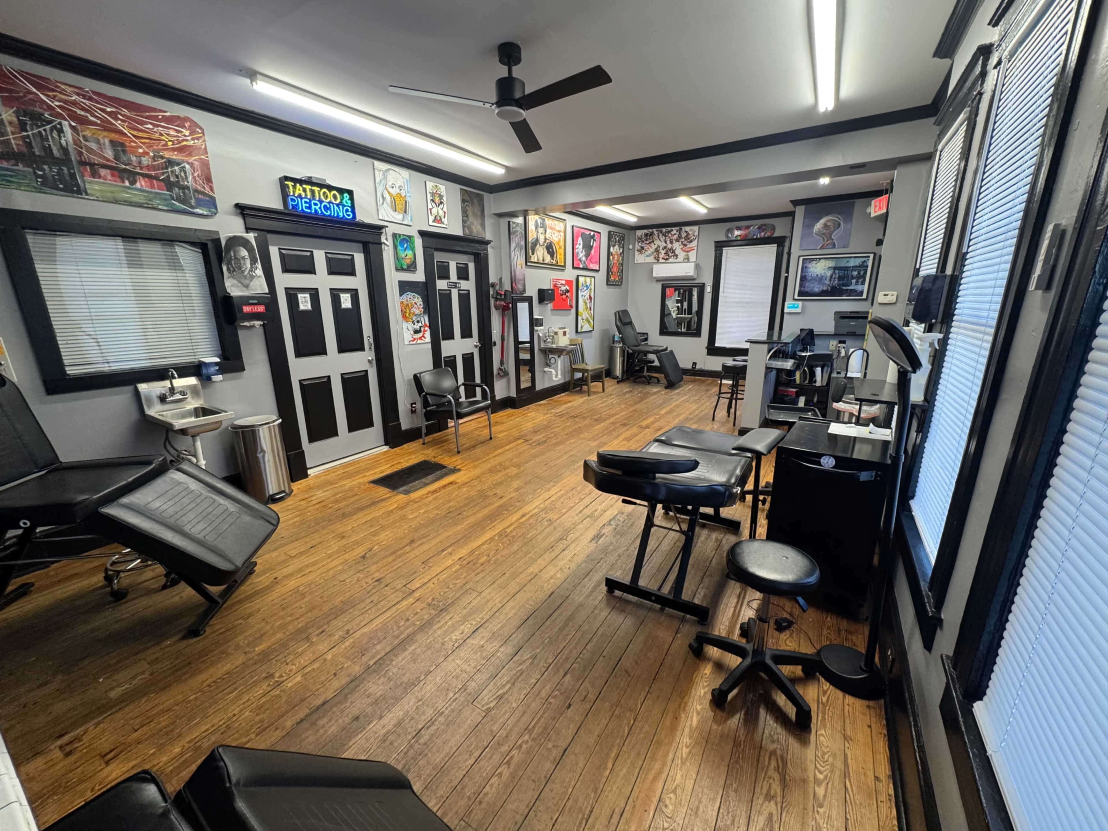 The image shows the interior of a tattoo studio featuring wooden flooring, several tattoo chairs, and a variety of artwork on the walls.