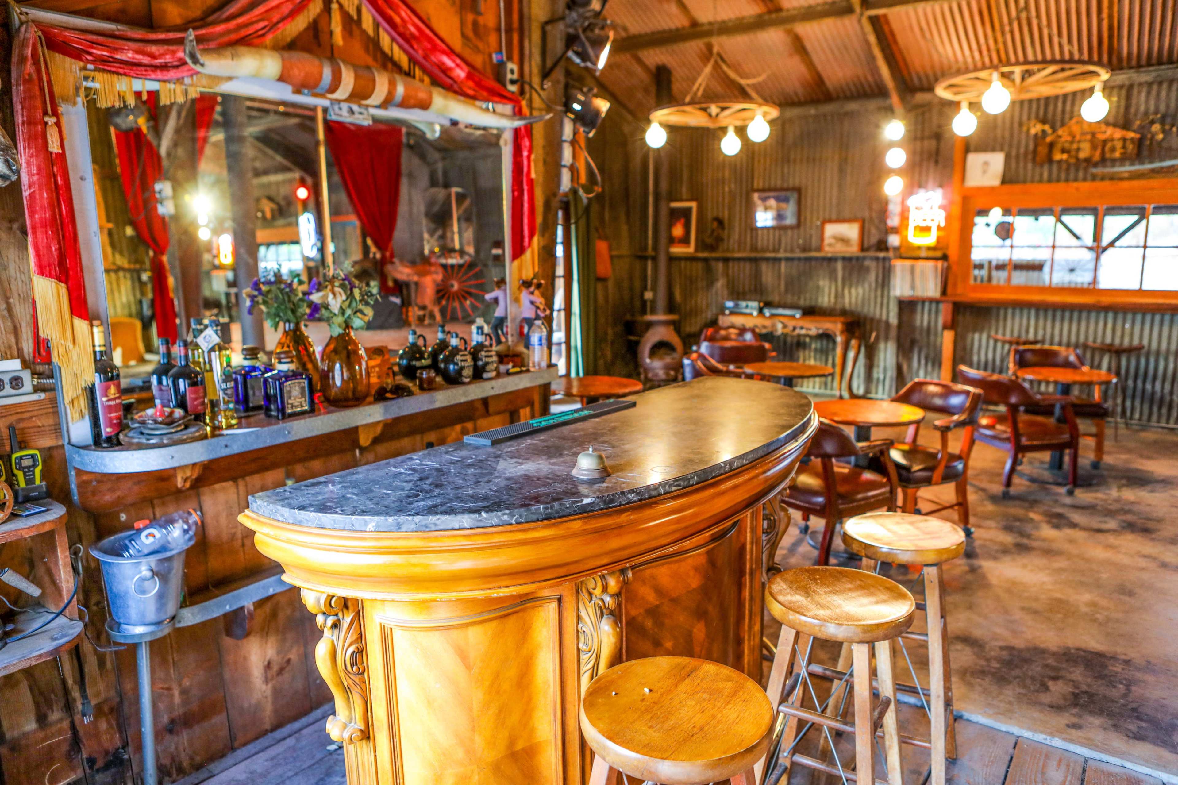 A rustic bar area with a curved wooden counter, high stools, and dim lighting reflecting off the corrugated metal walls.