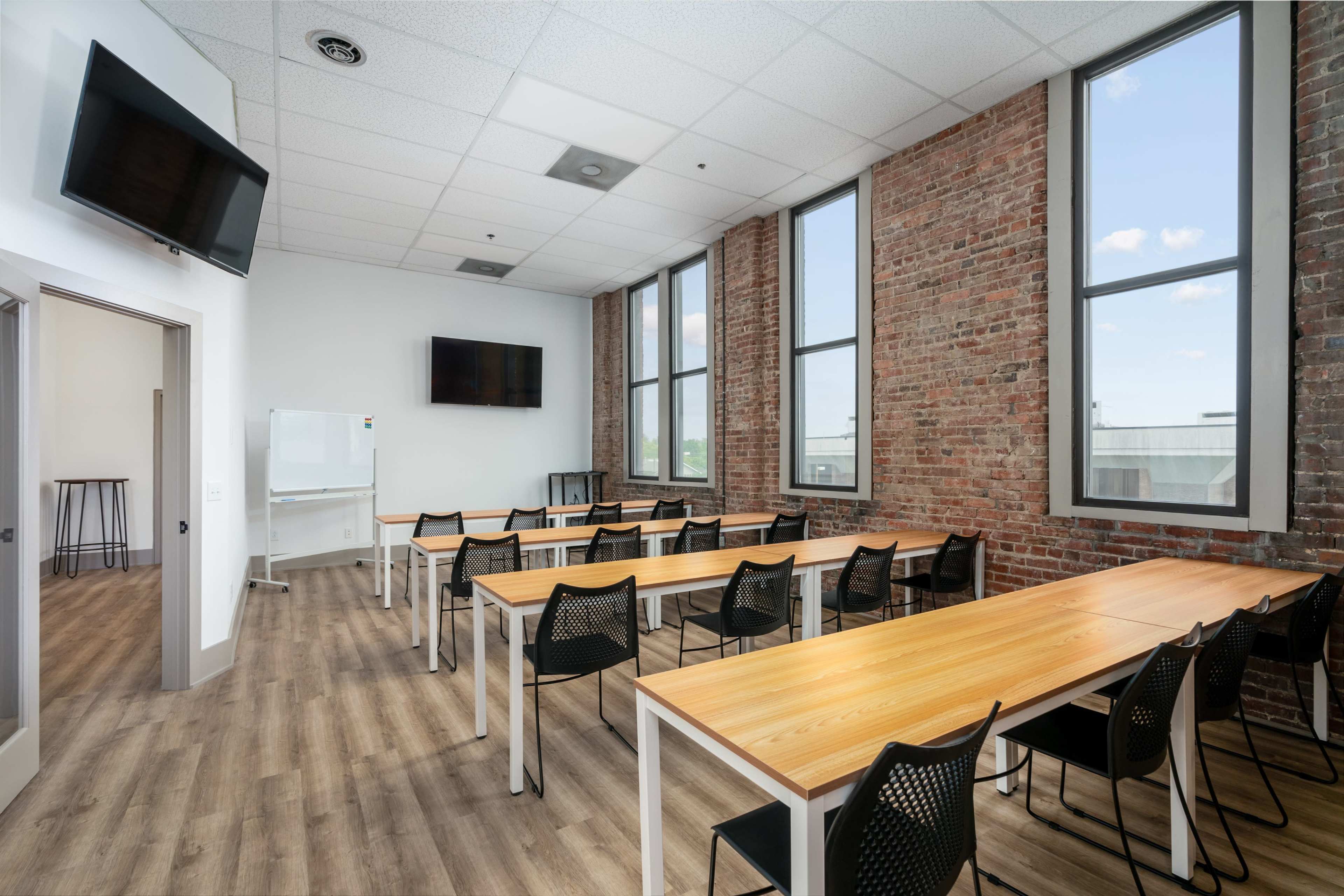 A modern classroom with wooden tables and black chairs, large windows, and exposed brick walls.