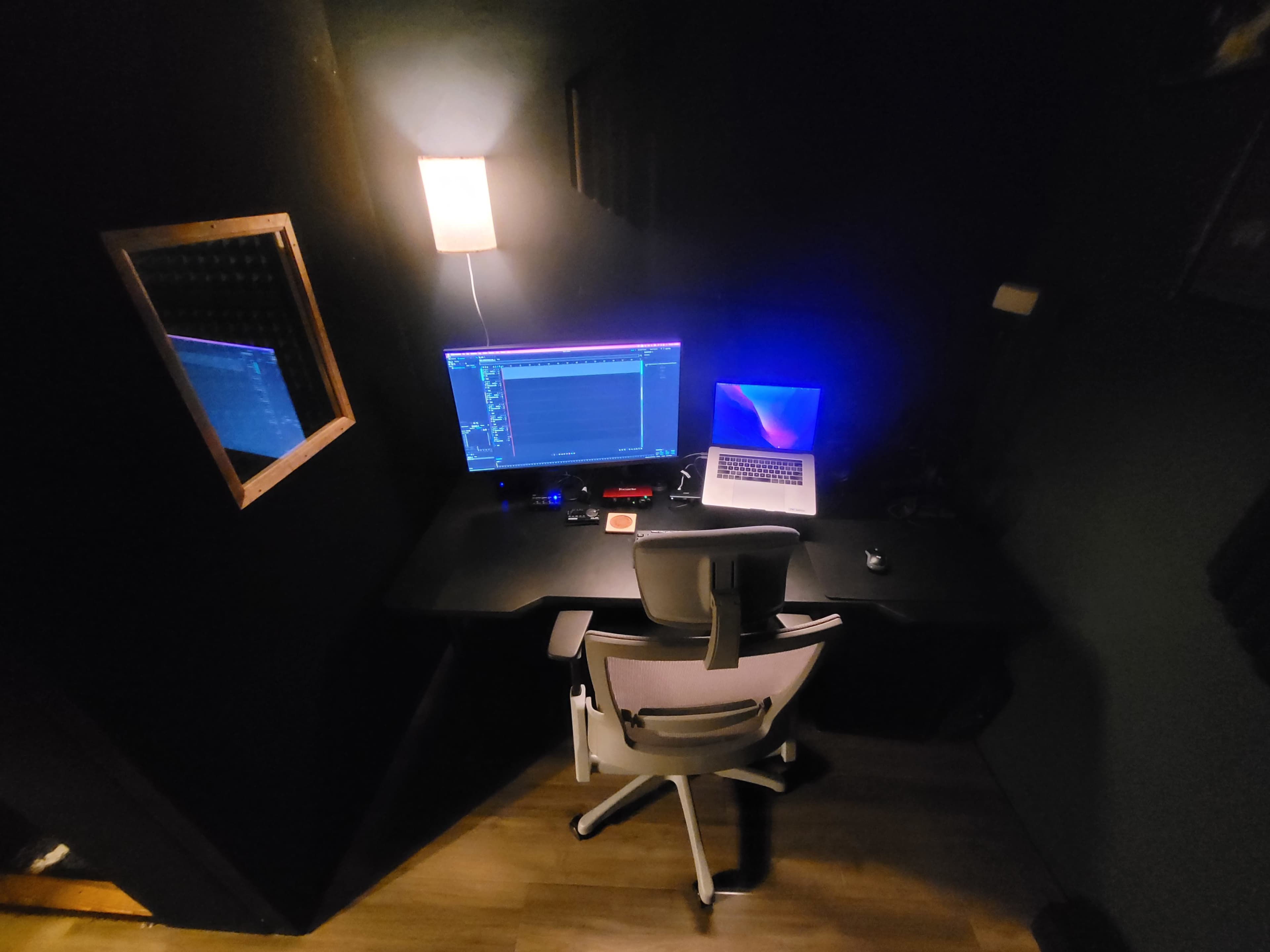 A dimly lit workspace features a desk with a large monitor, a laptop, and an ergonomic chair, complemented by a small wall mirror and a lamp.