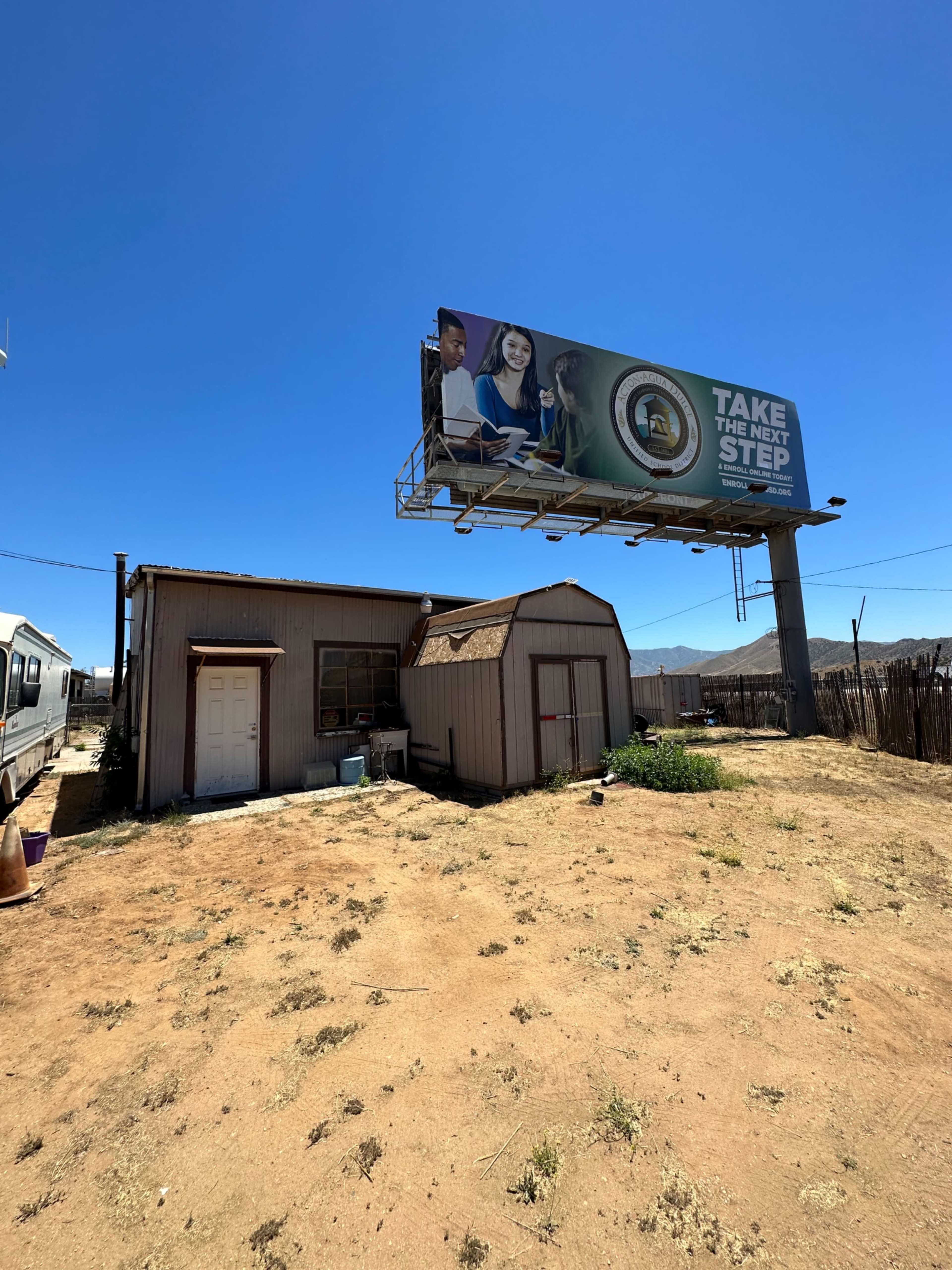 A small, weathered building with a metal roof sits in an arid landscape beneath a large billboard advertising a program.