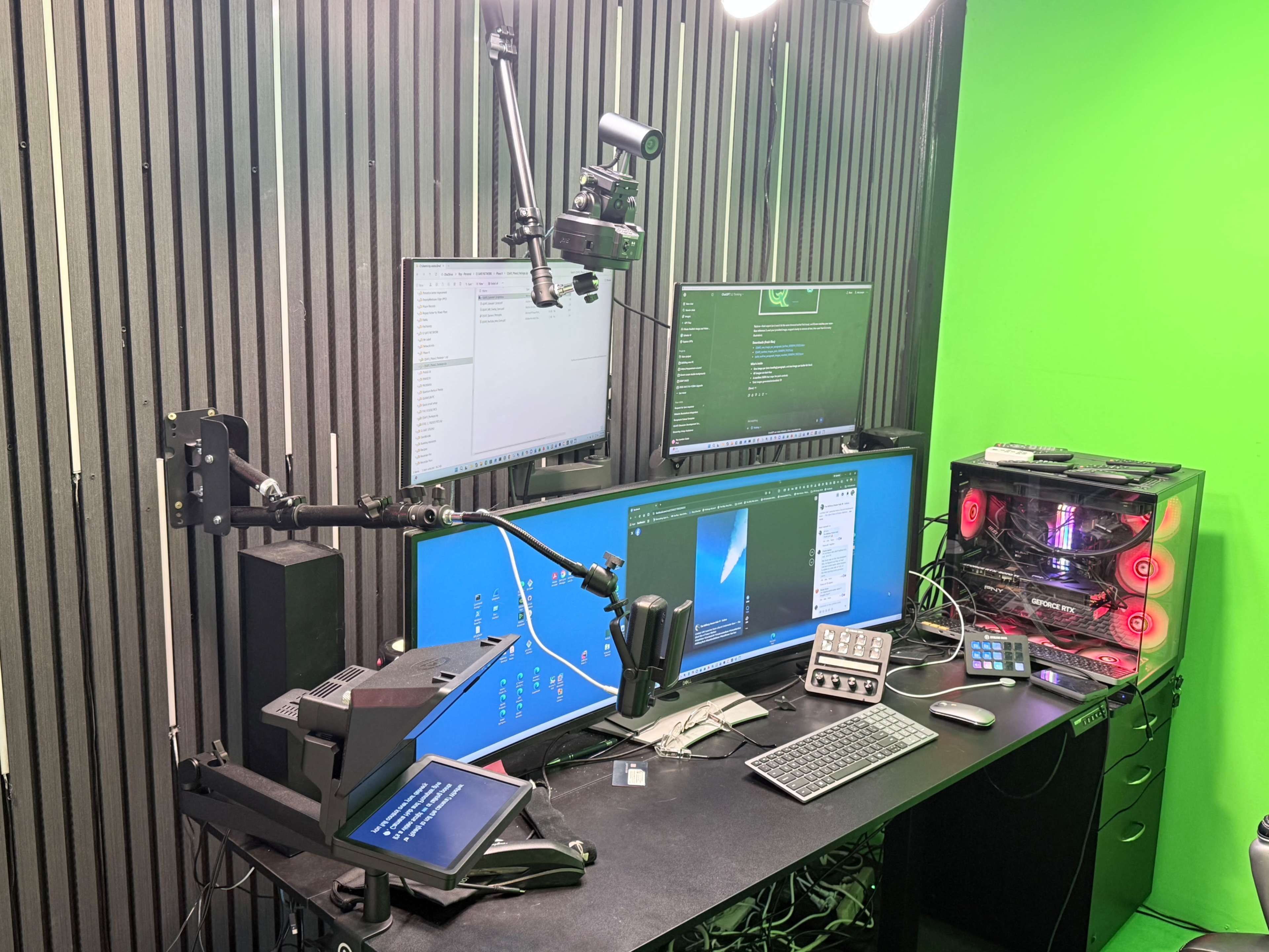 The image shows a modern workspace setup with multiple monitors, a camera mounted on an adjustable arm, a gaming PC with colored lighting, and various tech accessories on a black desk.
