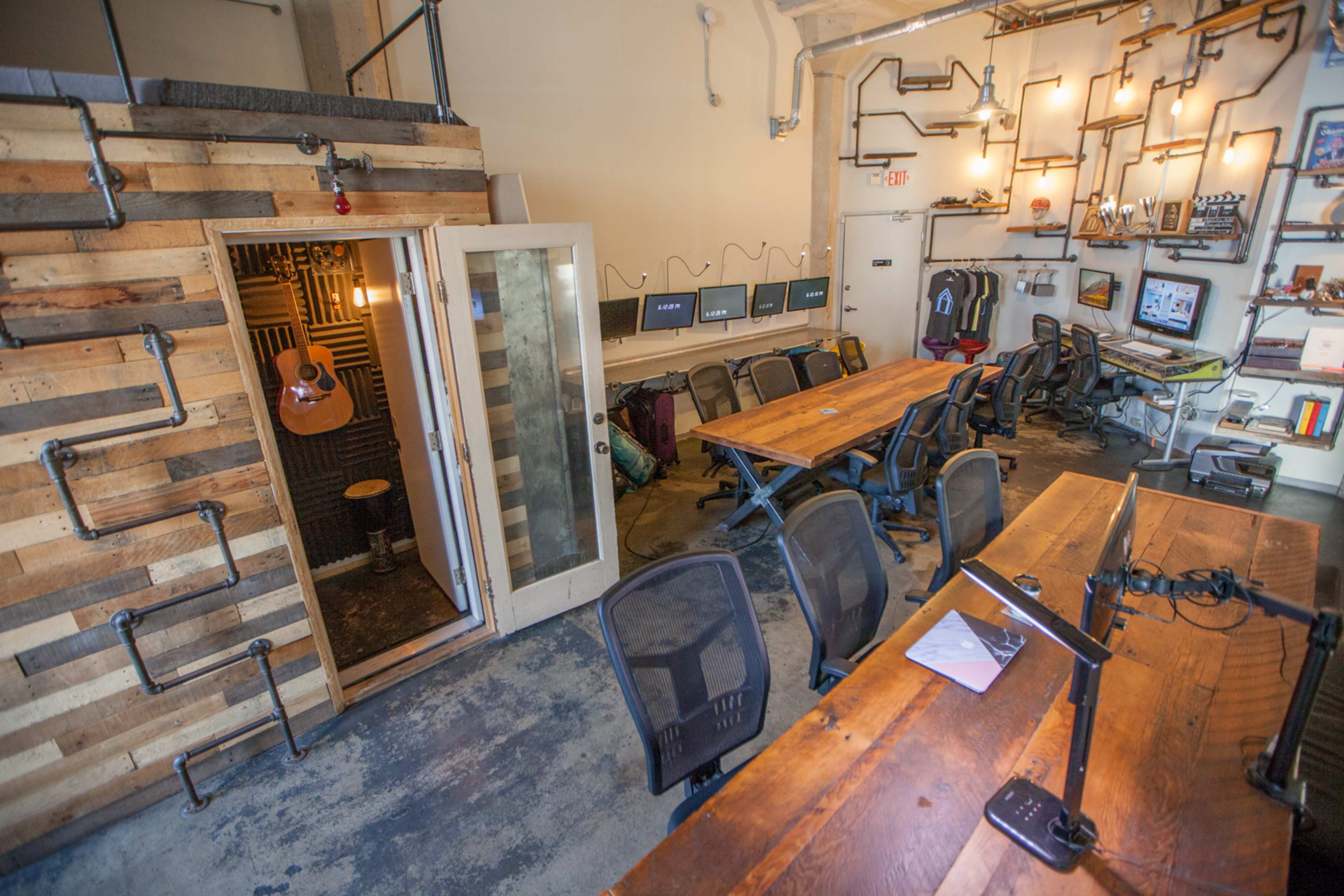 A modern workspace with a wooden conference table, office chairs, and a small soundproof room with an acoustic guitar inside.