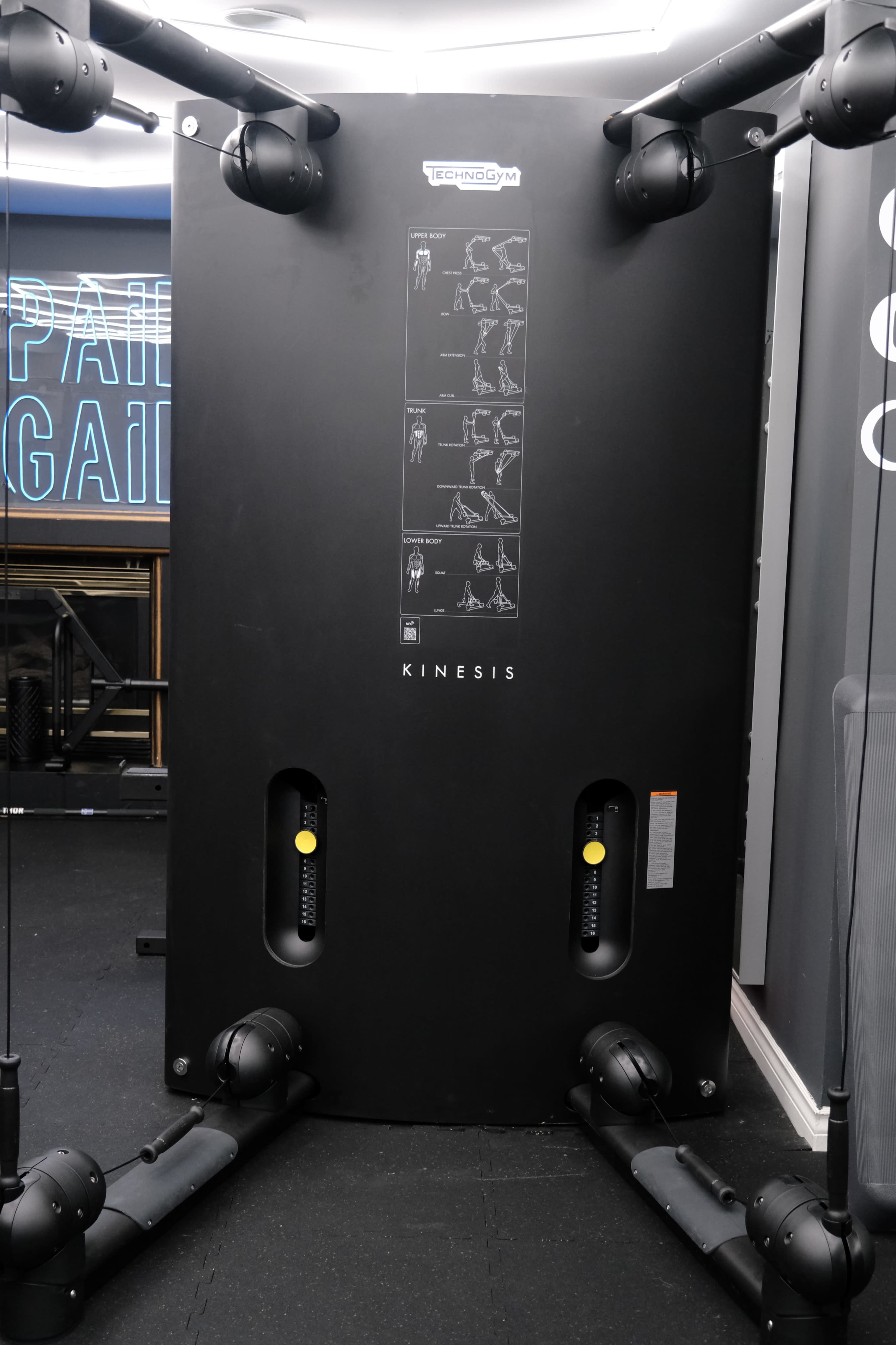 A black Kinesis gym machine featuring multiple pulleys and instructional diagrams for exercises.