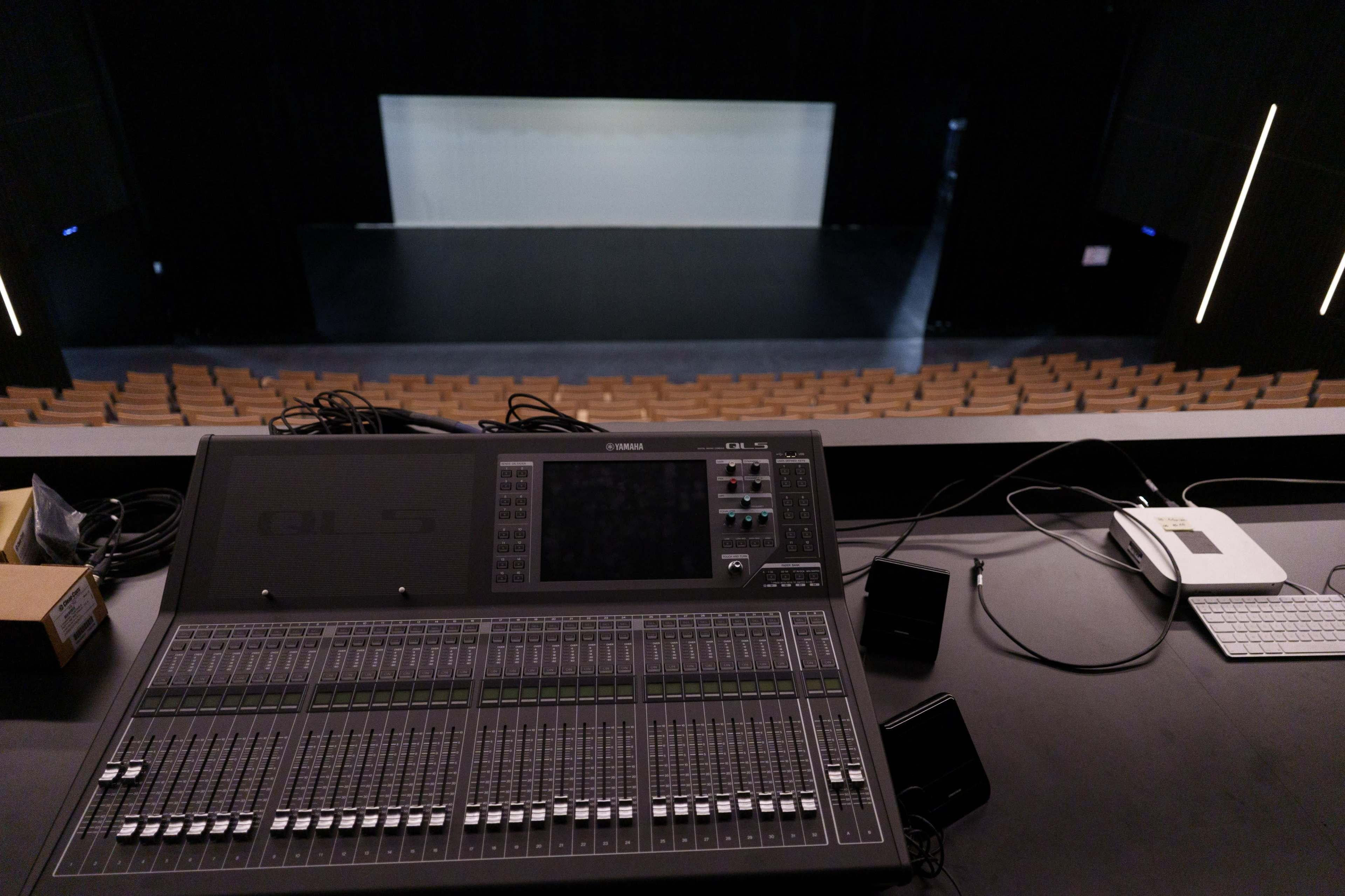 A sound mixing console is positioned in the foreground, overlooking a dimly lit theater stage with empty seating in the background.