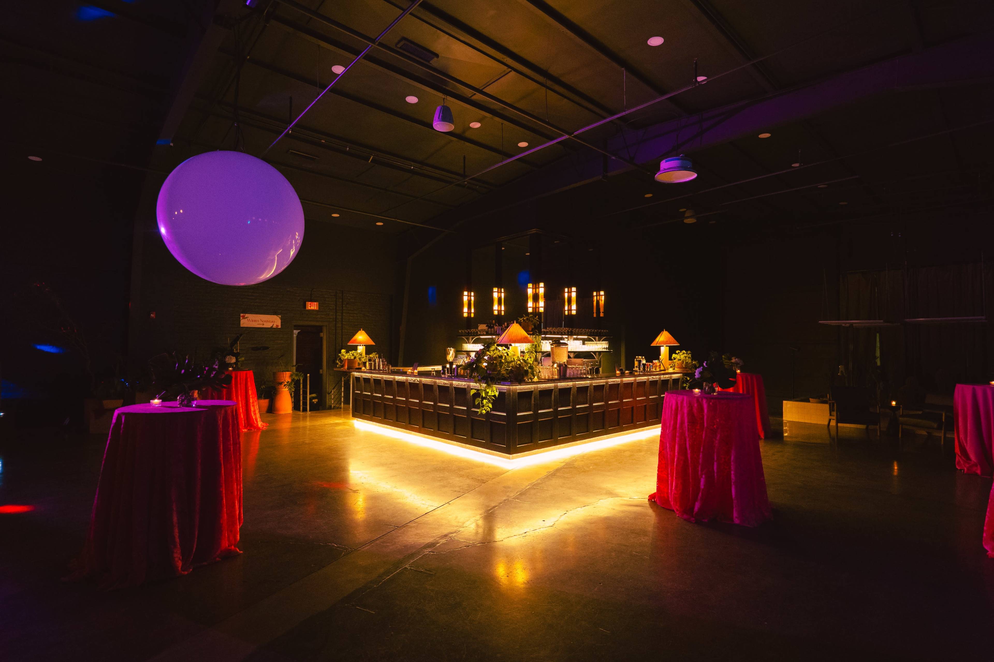 The image shows a dimly lit event space featuring a bar illuminated with warm lights, surrounded by tables dressed in pink tablecloths.