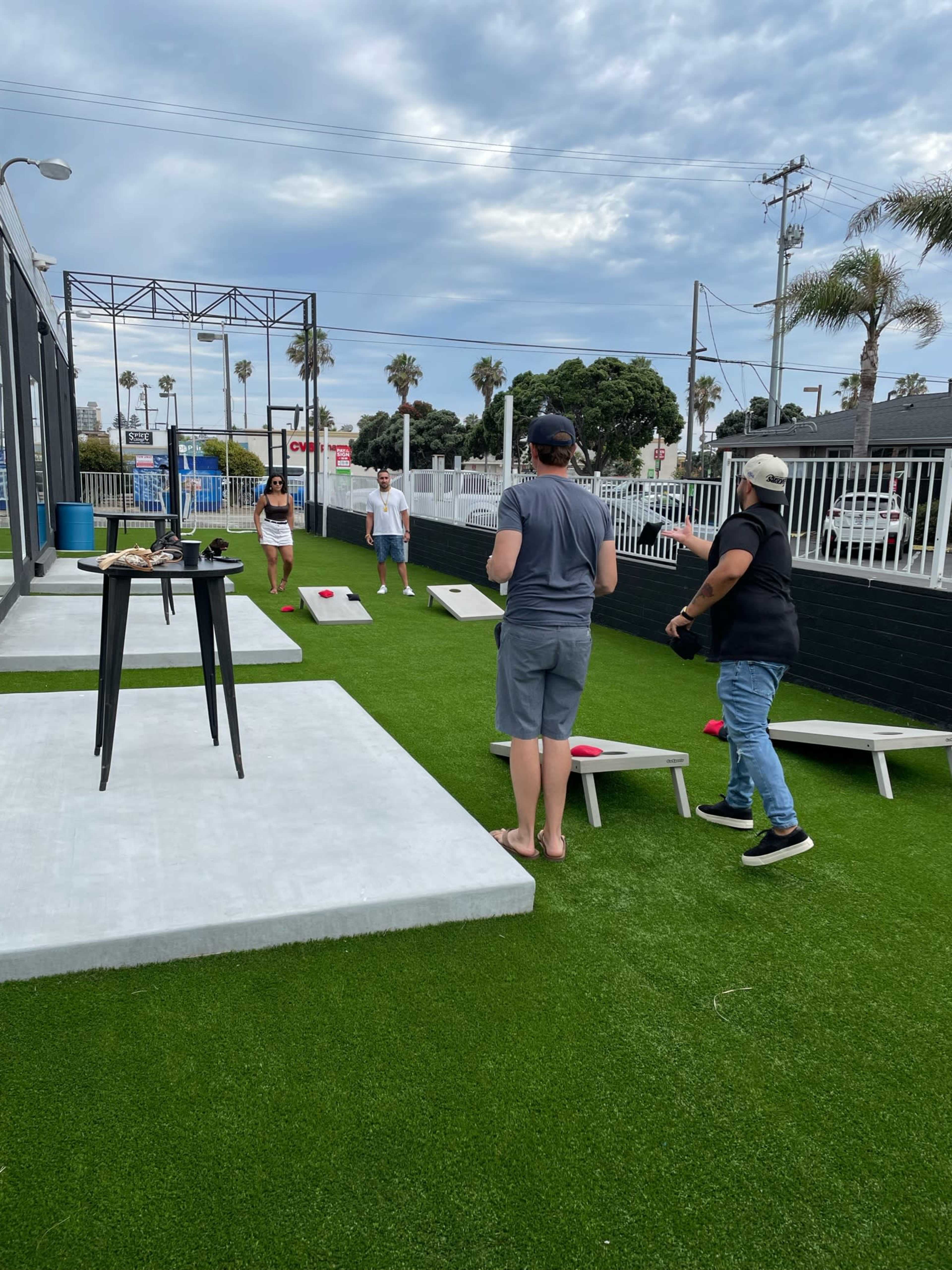 Four people play cornhole on a grassy outdoor area with palm trees in the background.