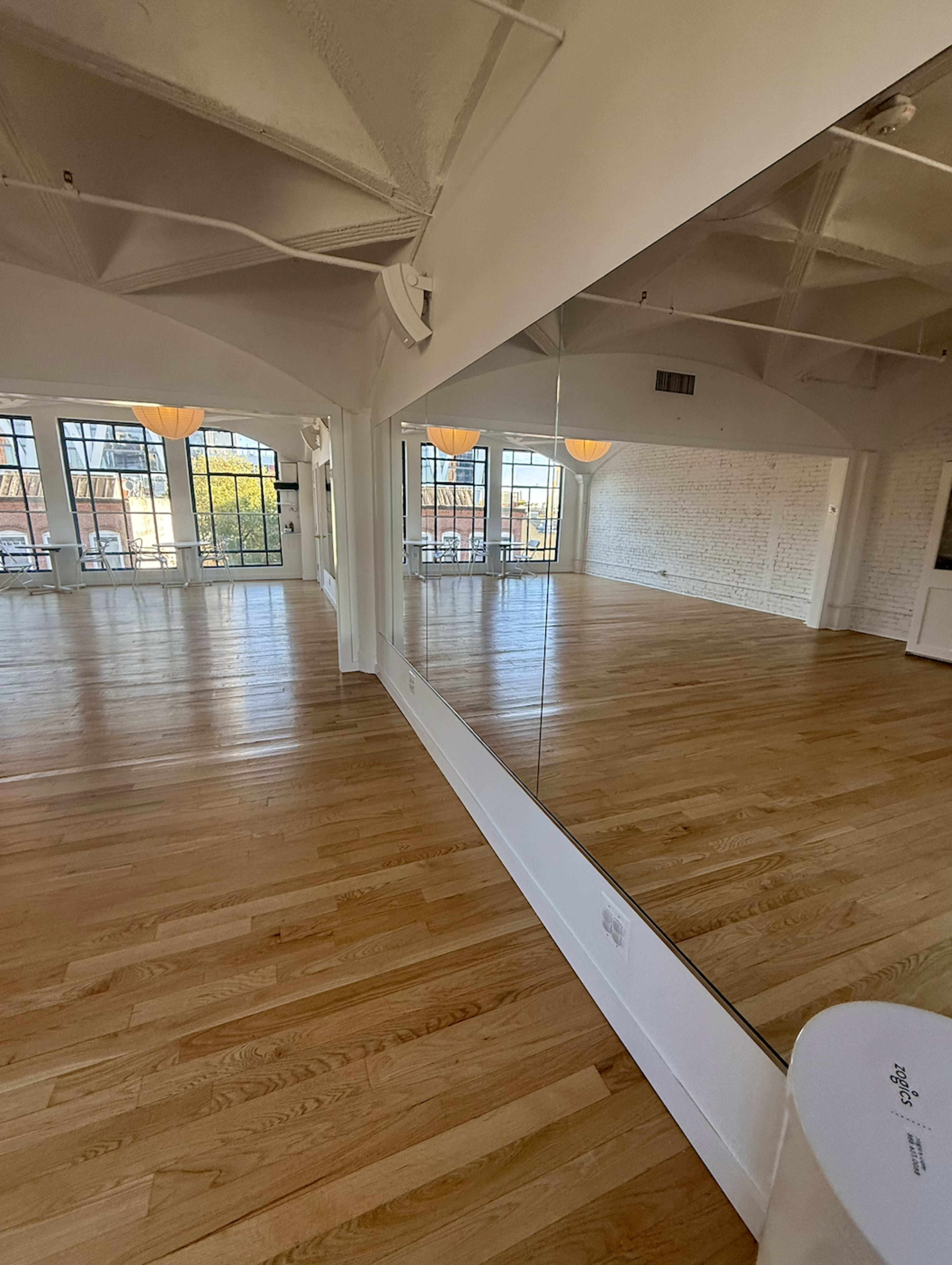 The image shows a spacious interior room with hardwood floors, large windows, and a mirrored wall.