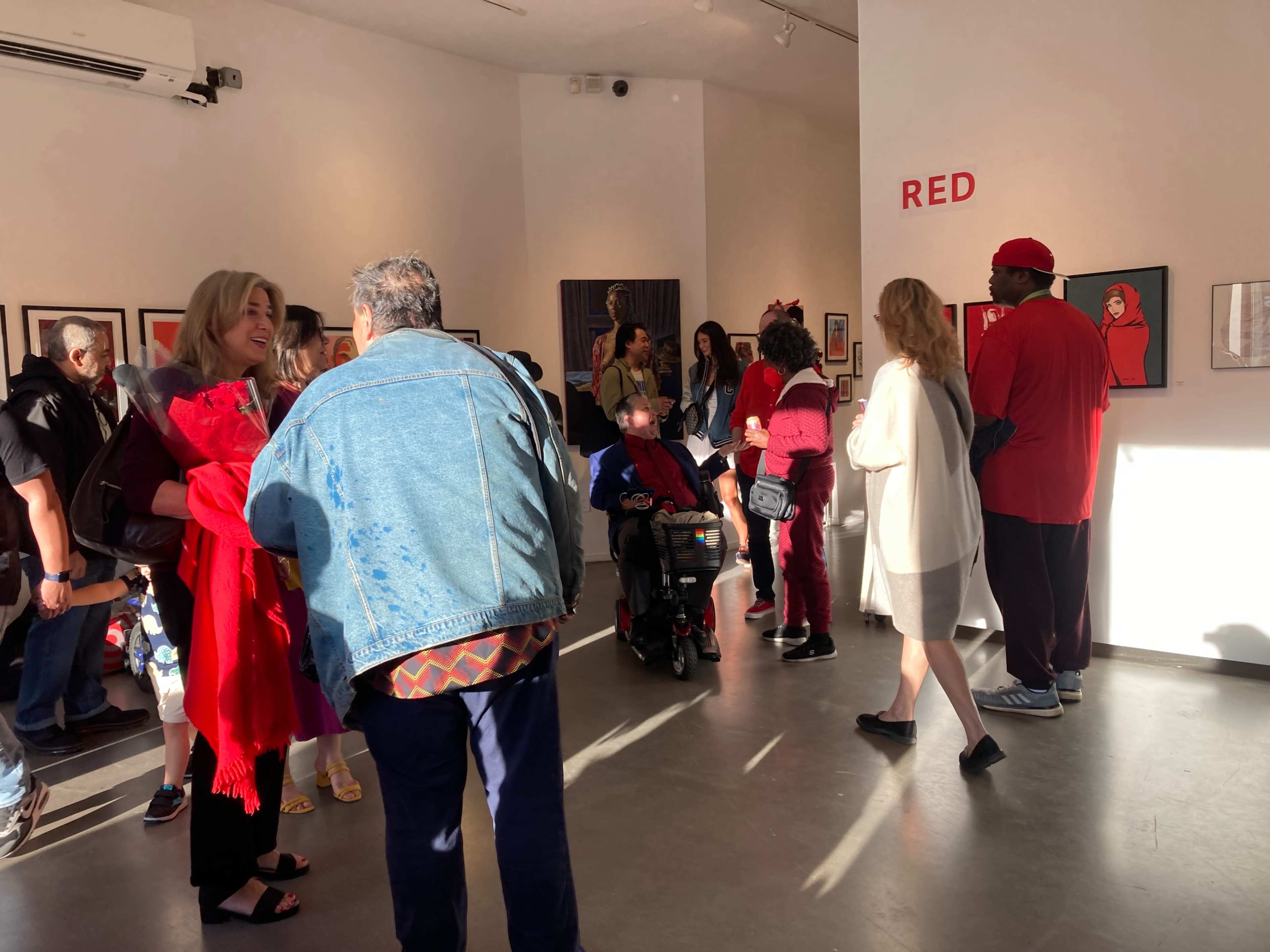 The image shows a gallery event with attendees in various outfits engaging and mingling around artwork on display.