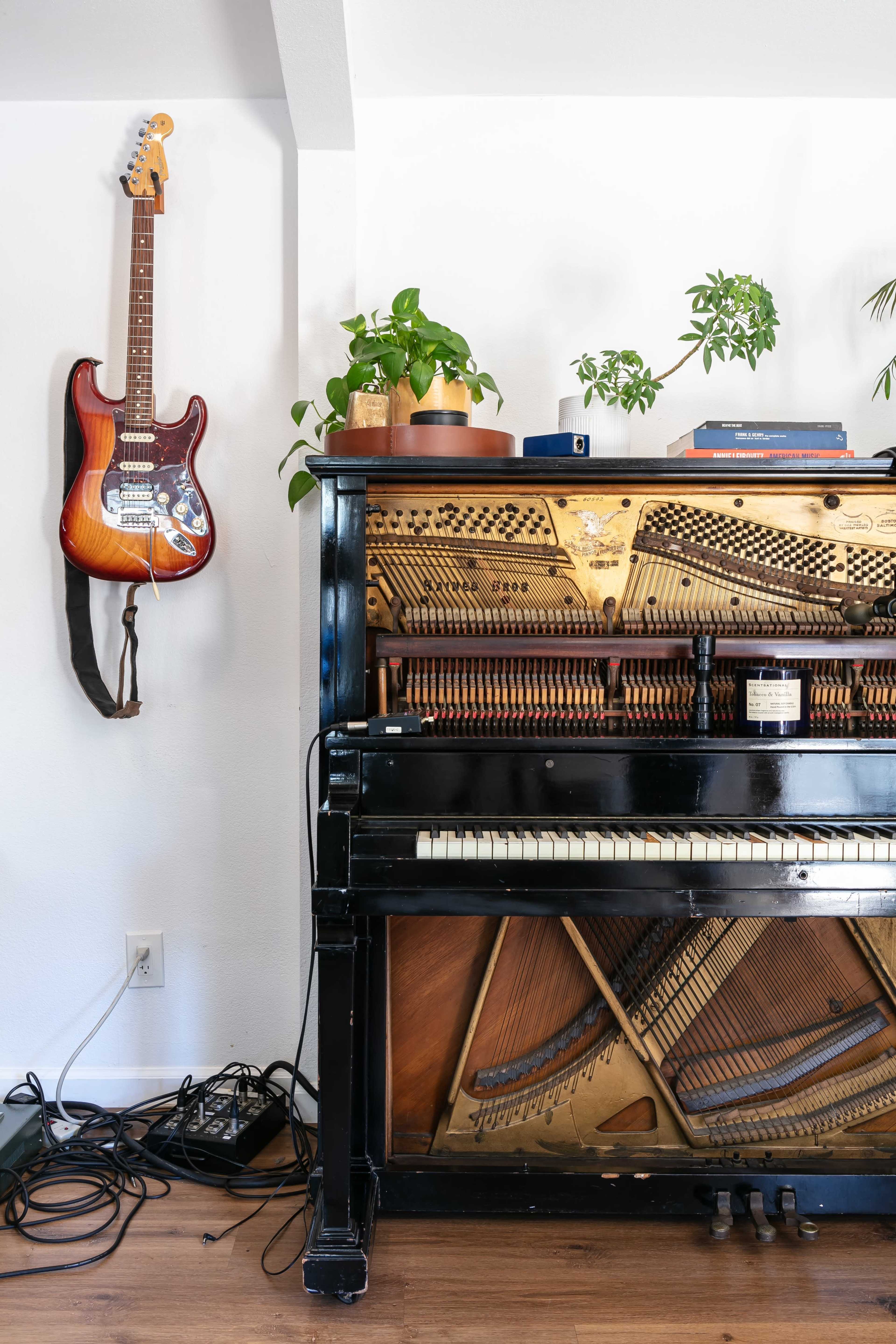 A black piano with exposed internal mechanisms stands beside a wall-mounted electric guitar and potted plants.