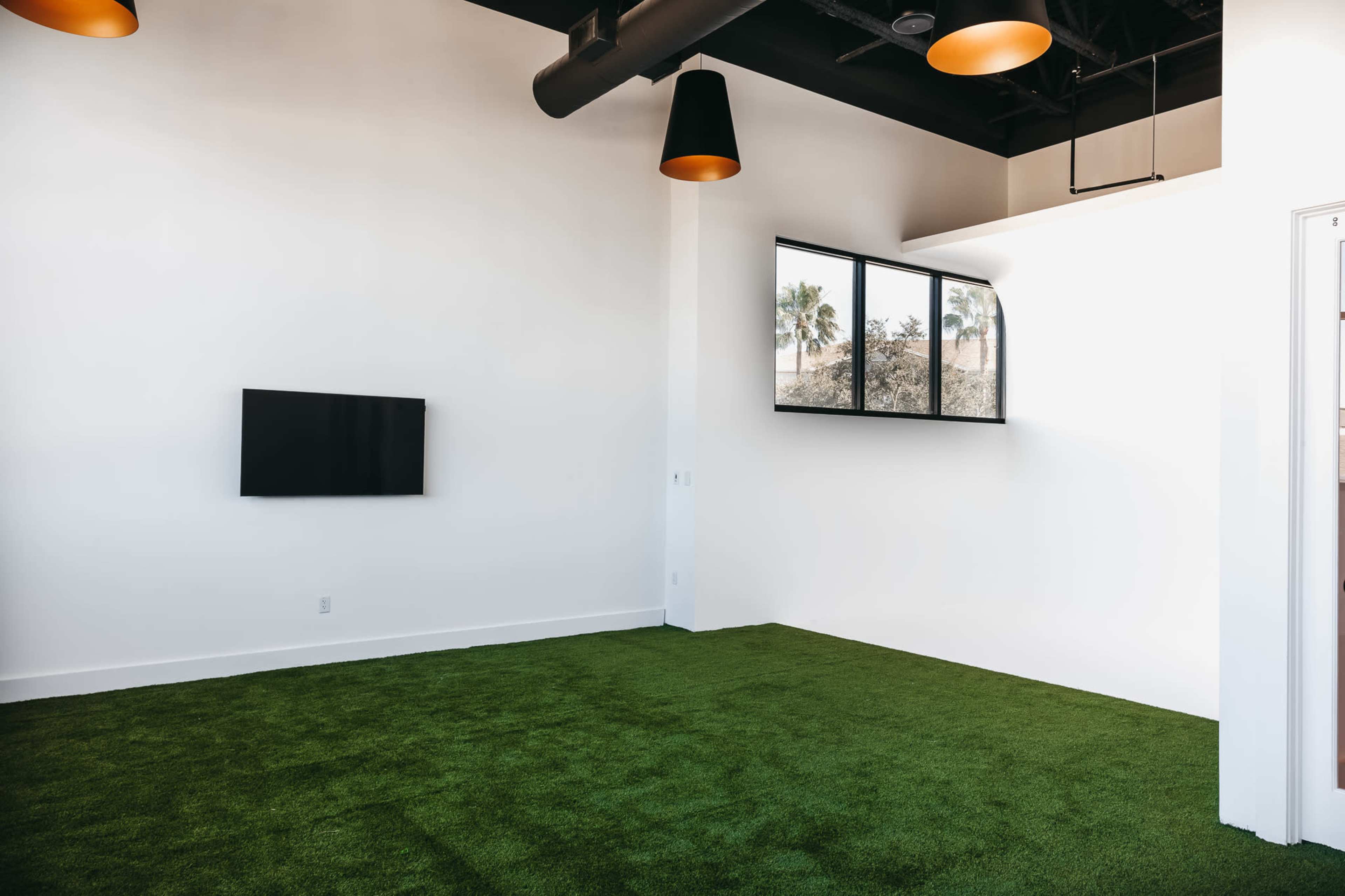 The image shows a minimalistic room with a green carpet, a flat-screen TV mounted on the white wall, and large windows allowing natural light to enter.