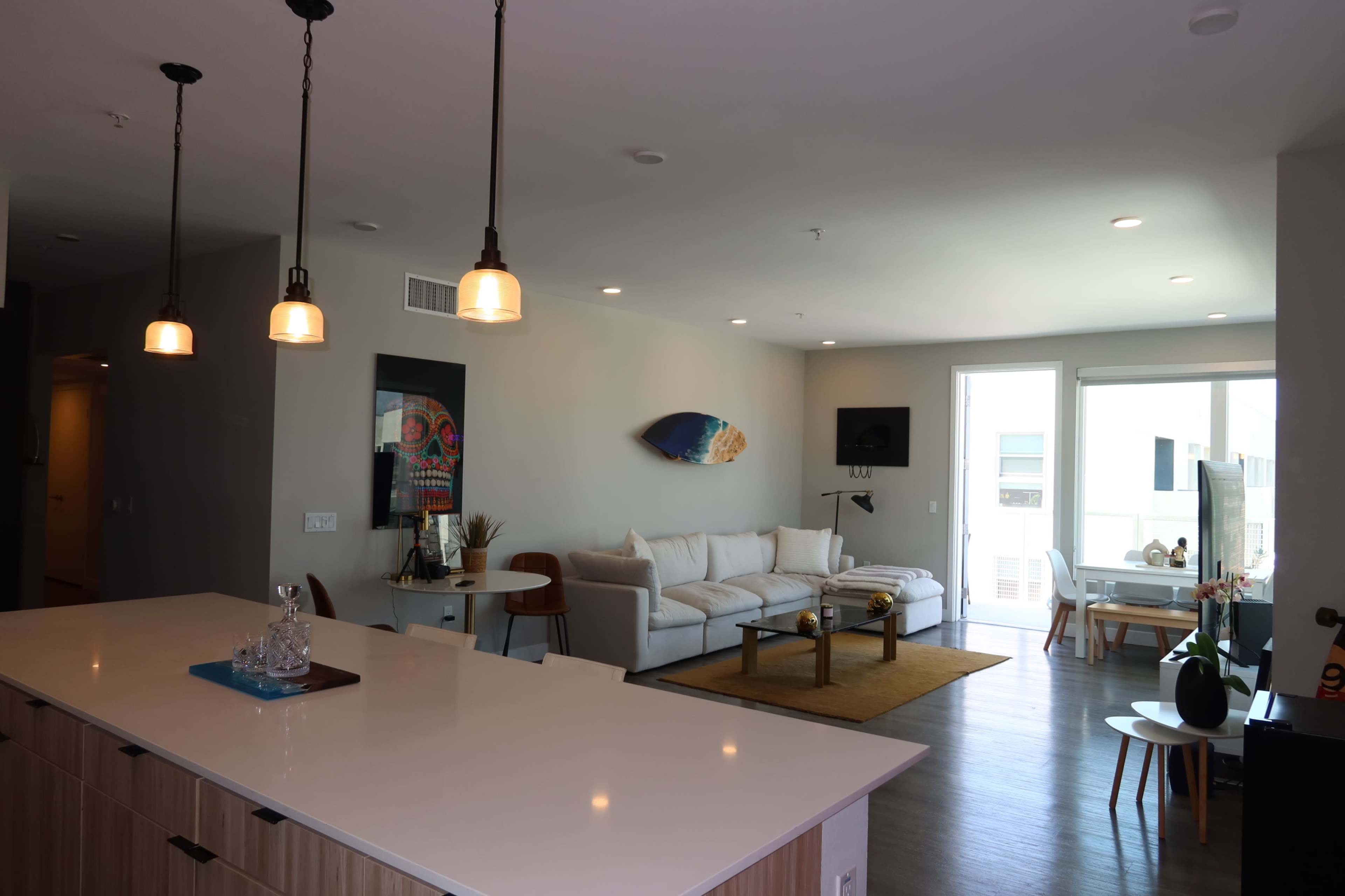 The image shows a modern living space featuring a white sofa, a dining area with bar stools, and large windows providing natural light.
