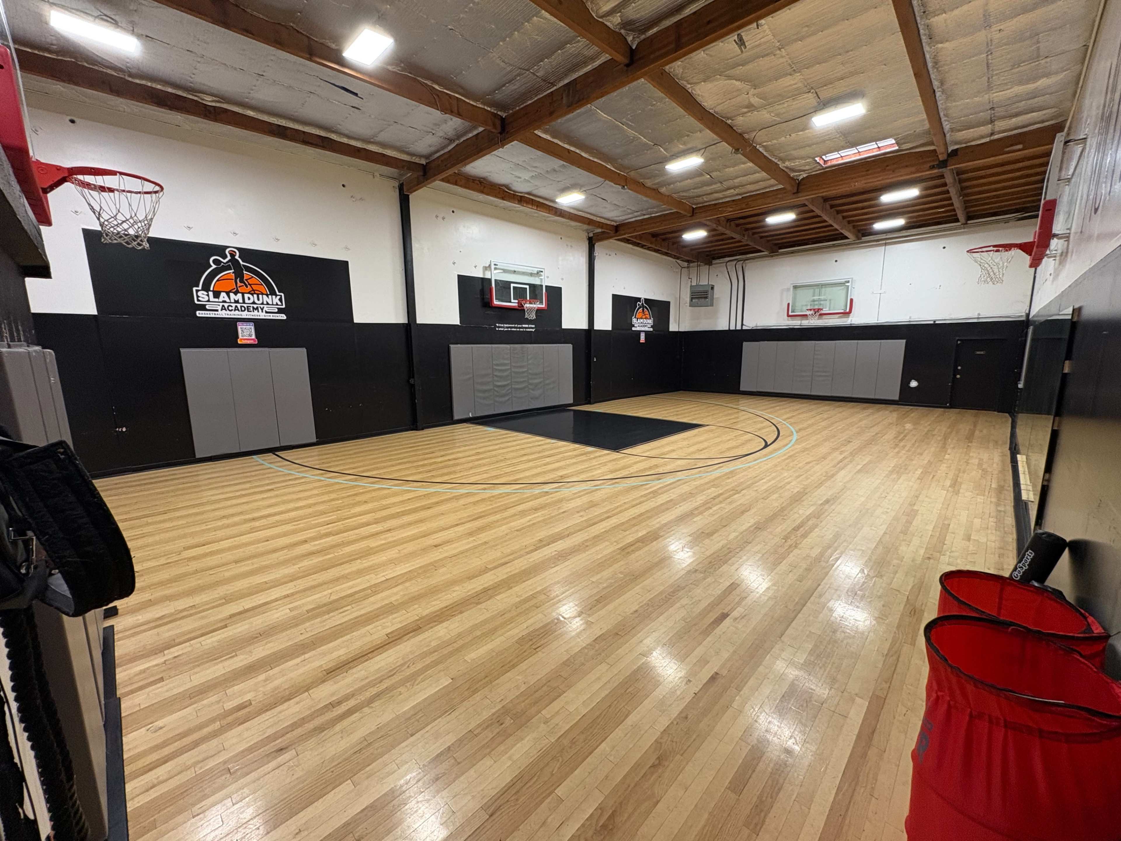 The image shows a basketball court with wooden flooring, two hoops at each end, and a logo on the wall.