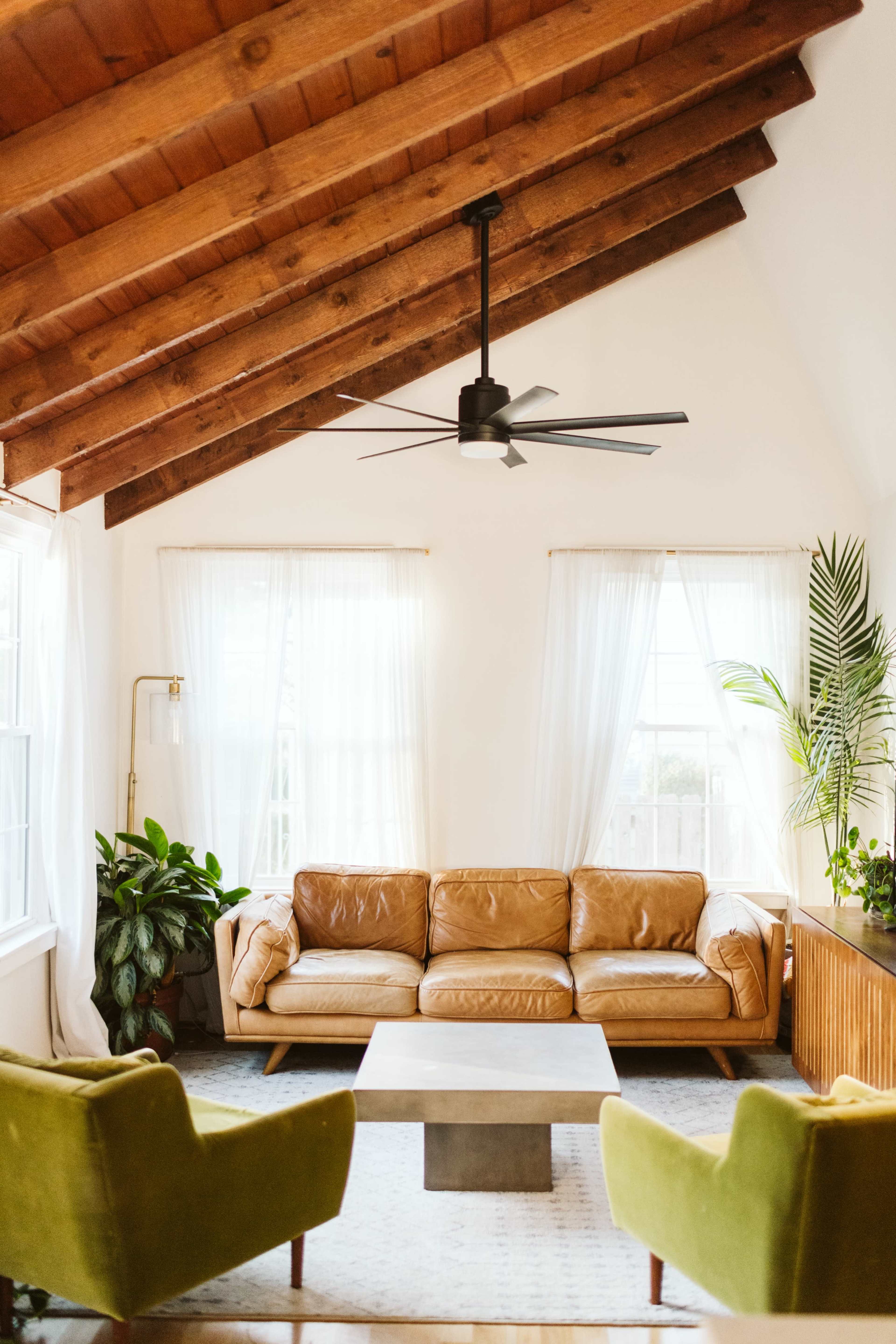 A cozy living room features a leather sofa, two green armchairs, a modern coffee table, and large windows with sheer curtains.