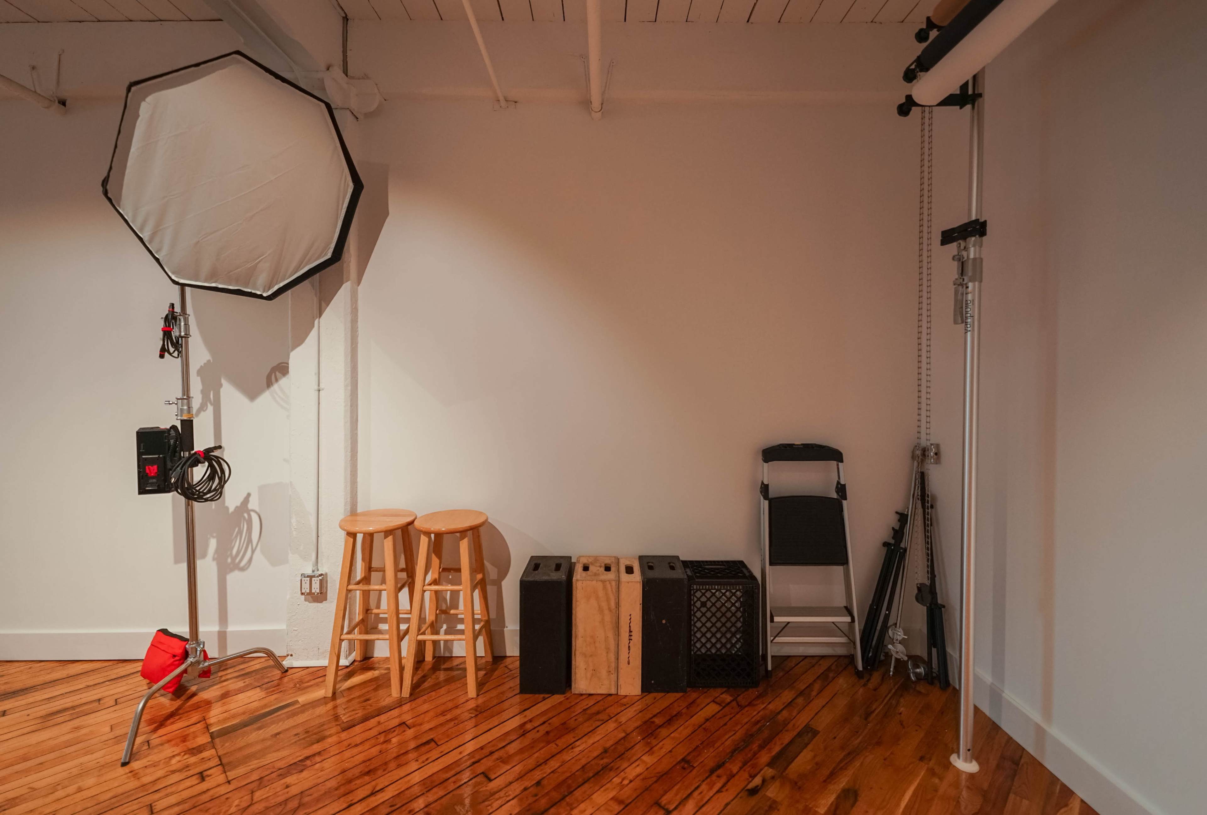 A minimalist photo studio with two wooden stools, various equipment boxes, and a lighting setup against a plain wall.