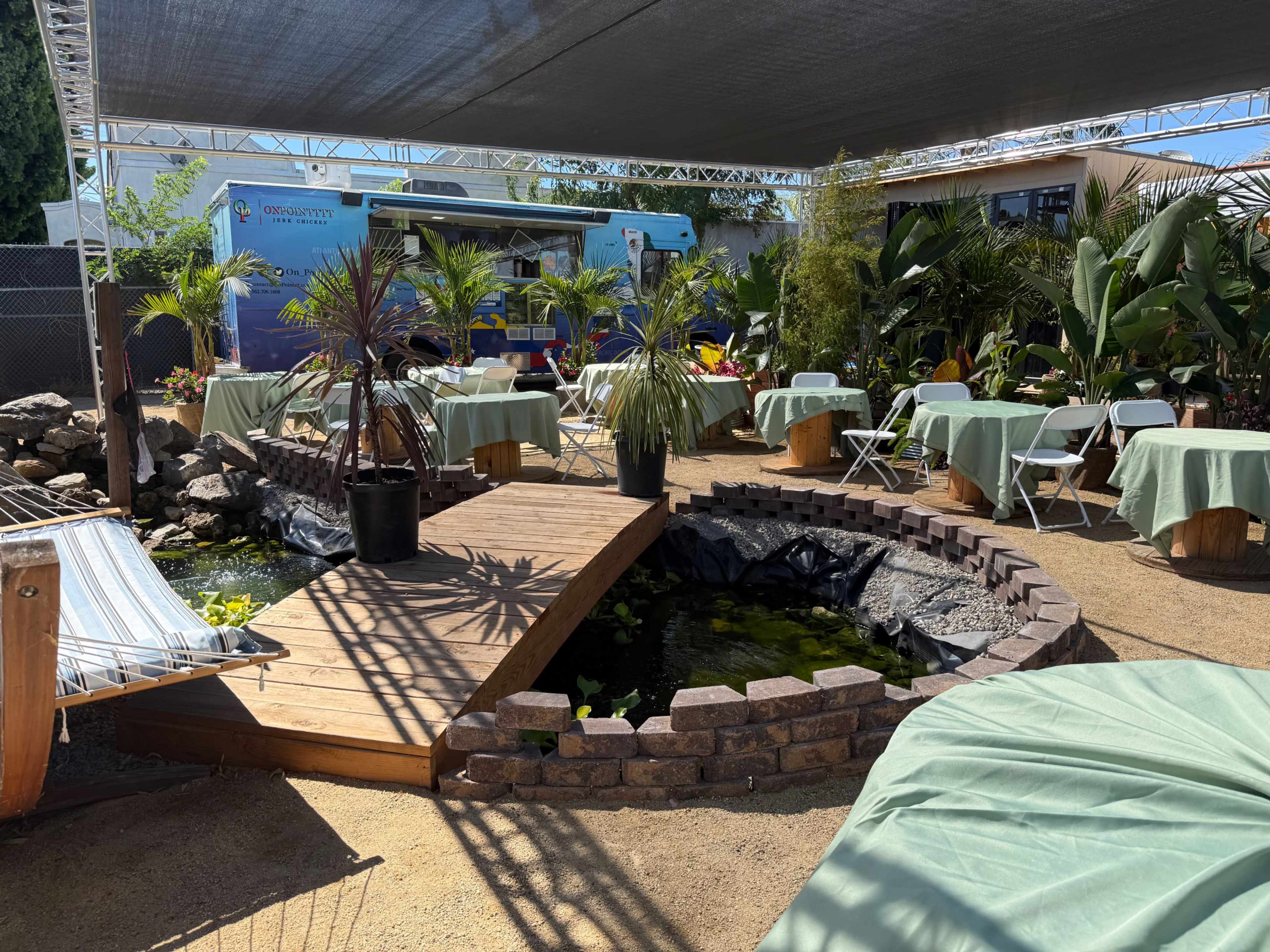 The image shows an outdoor dining area featuring bamboo seating, green tables covered with fabric, a wooden bridge over a pond, and decorative plants.