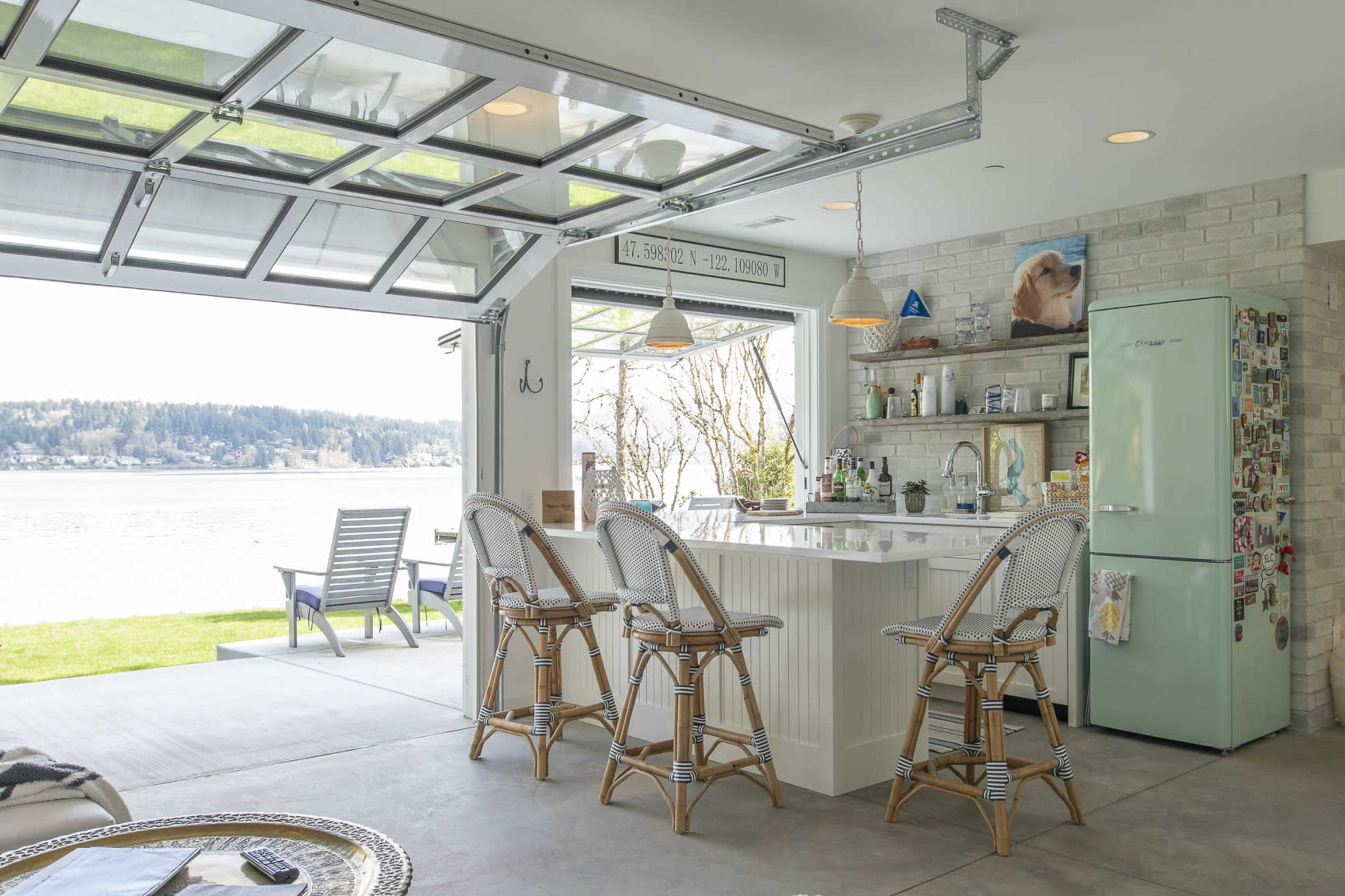 A modern kitchen with a bar and four rattan stools opens to a scenic view of a body of water through large glass doors.