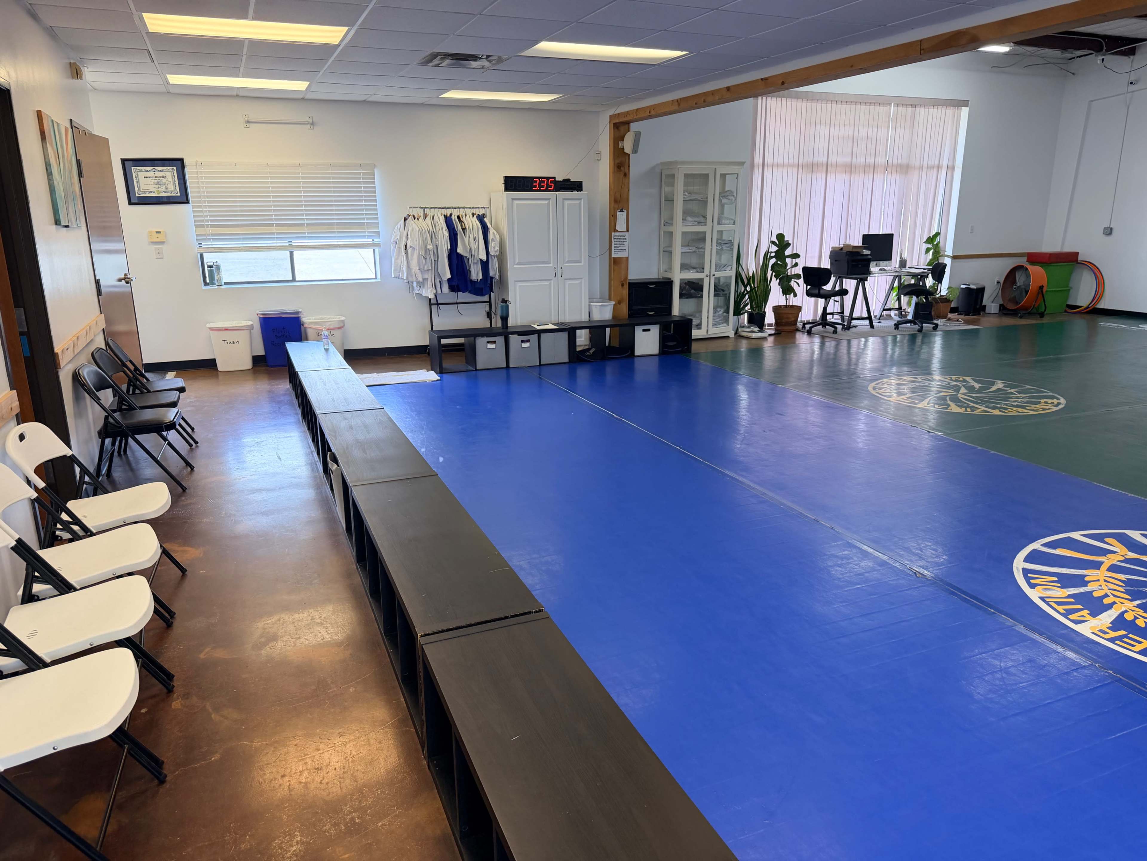 The image shows a spacious martial arts training room with blue and green mats, a bench for seating, and equipment organized in the background.