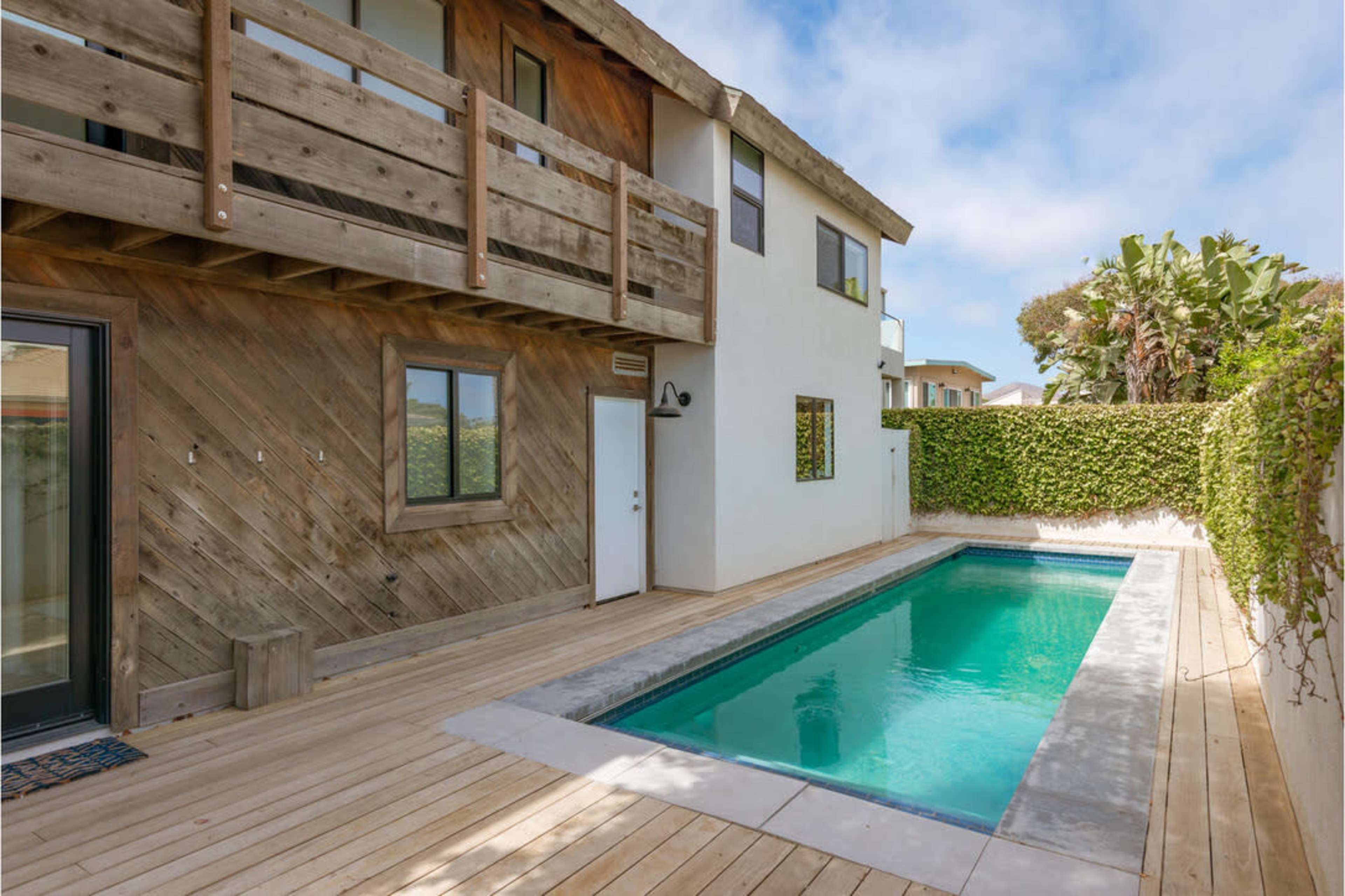 A modern house with a wooden deck and a rectangular swimming pool surrounded by greenery.