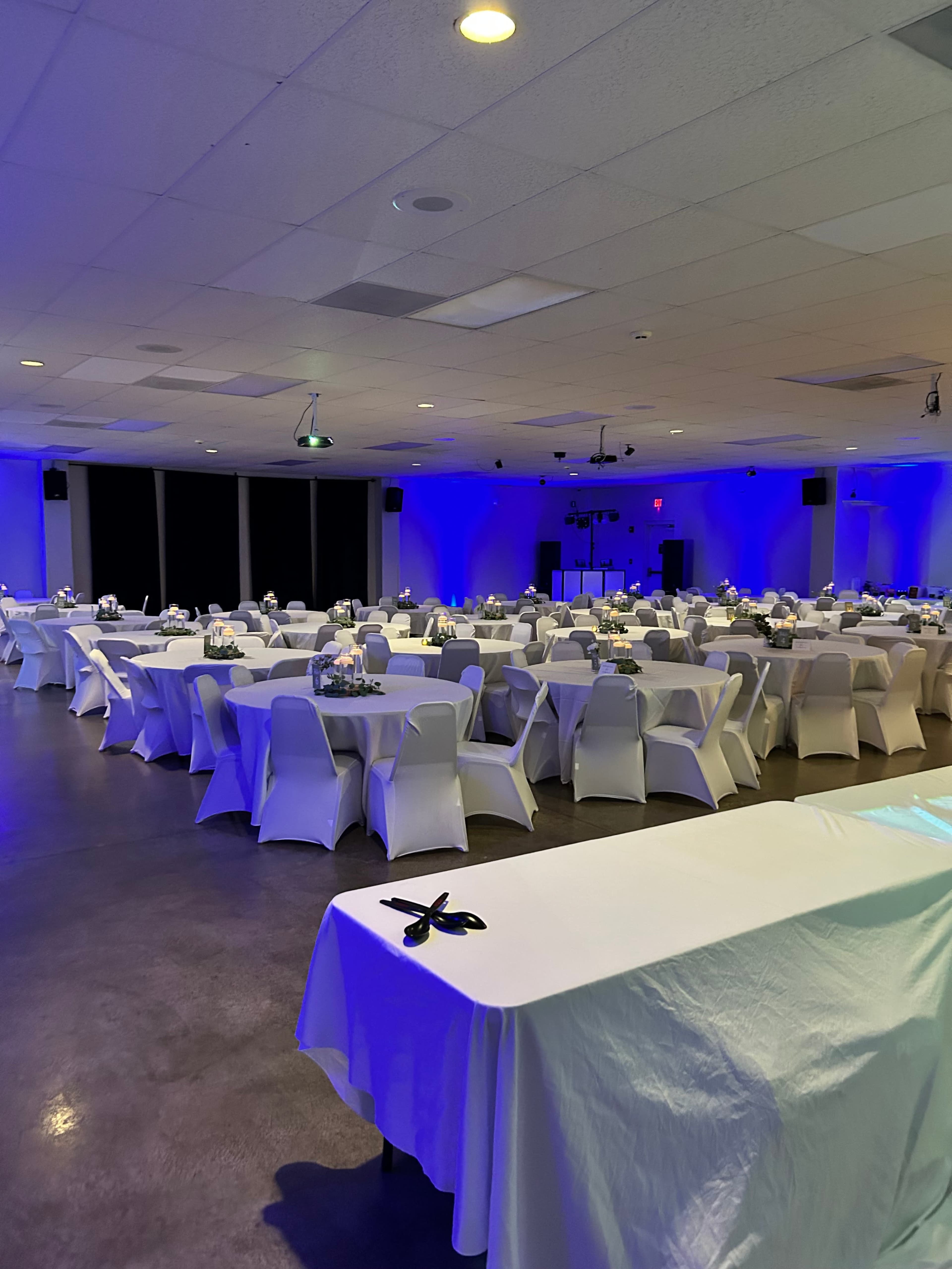 A large event space is set with rows of white tables covered with cloths and decorated with floral centerpieces under blue lighting.