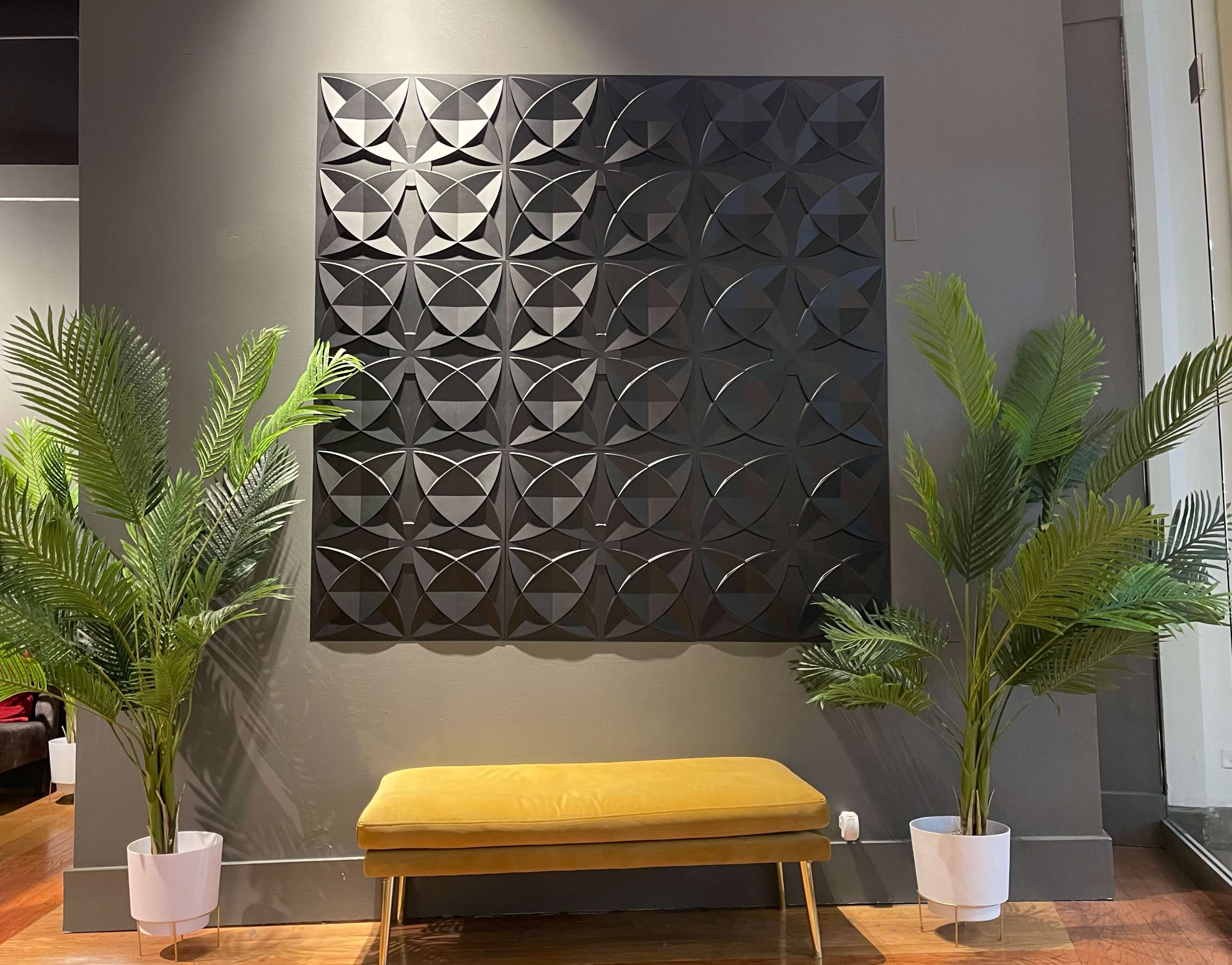 A textured black wall panel is centered above a mustard yellow bench, flanked by two potted palm plants.