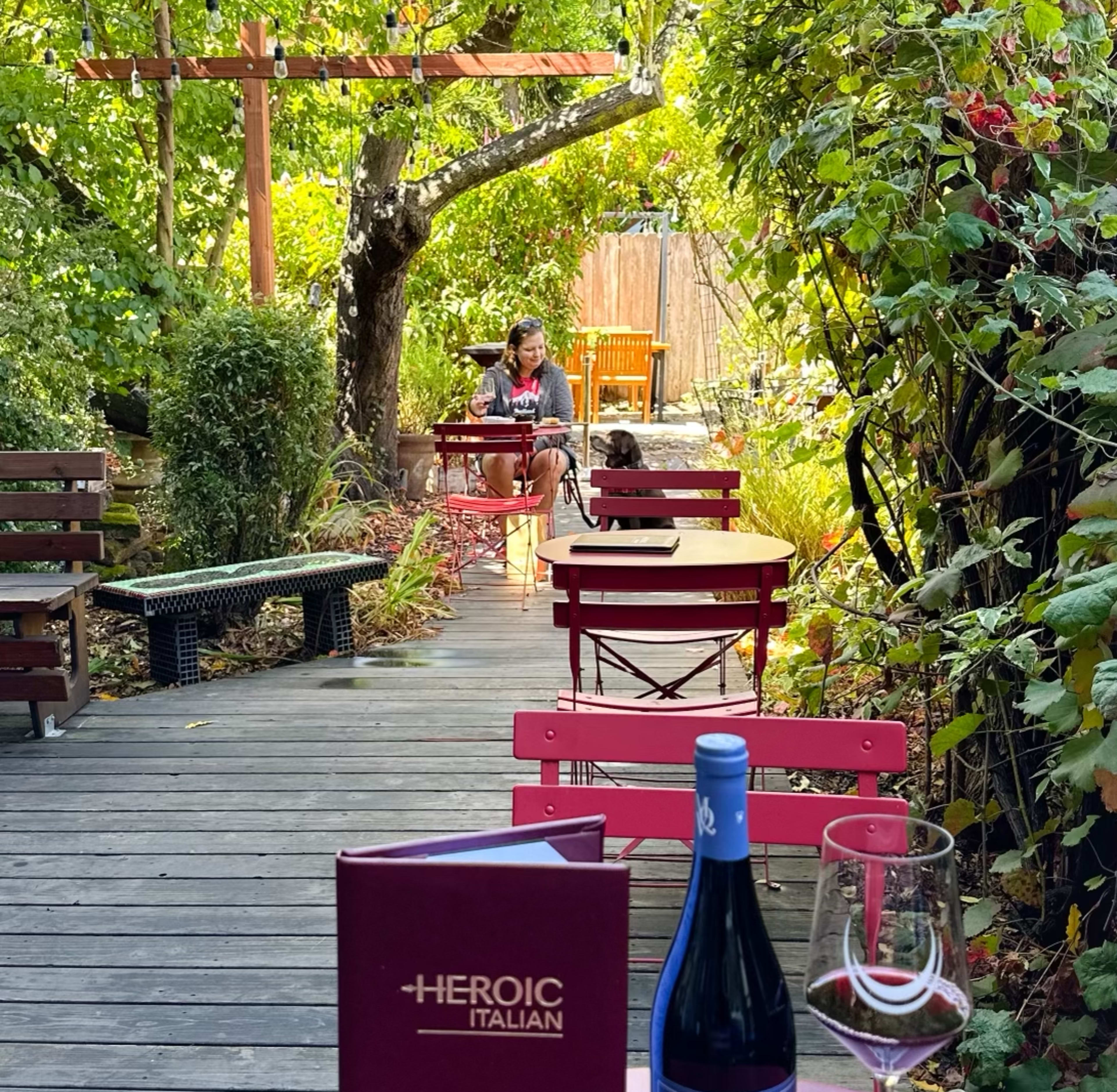A winding wooden pathway in a garden features red chairs and a nearby table with a wine glass and menu, while a person sits in the distance with a dog.