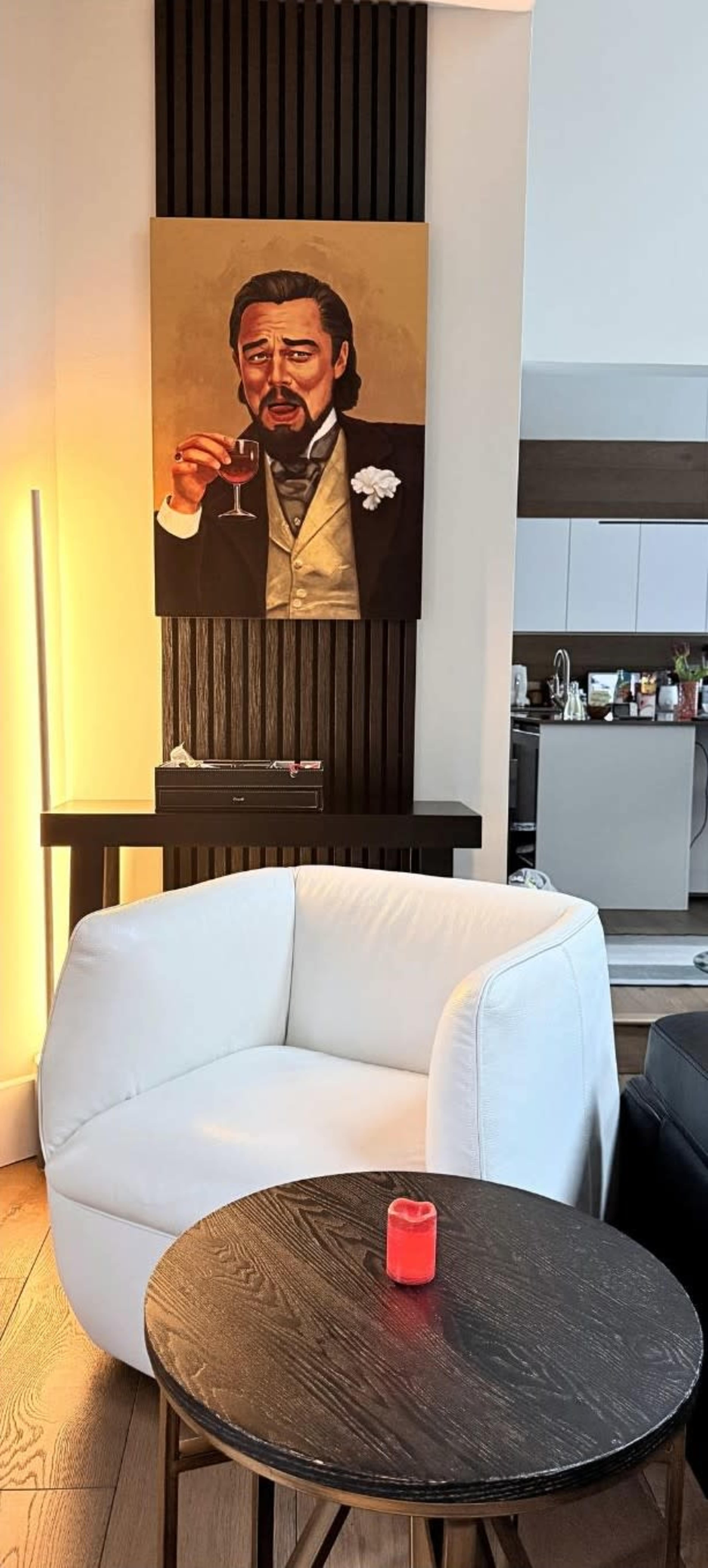 The image shows a modern interior with a white chair and a black side table featuring a candle, while a portrait of a man holding a glass appears on the wall in the background.