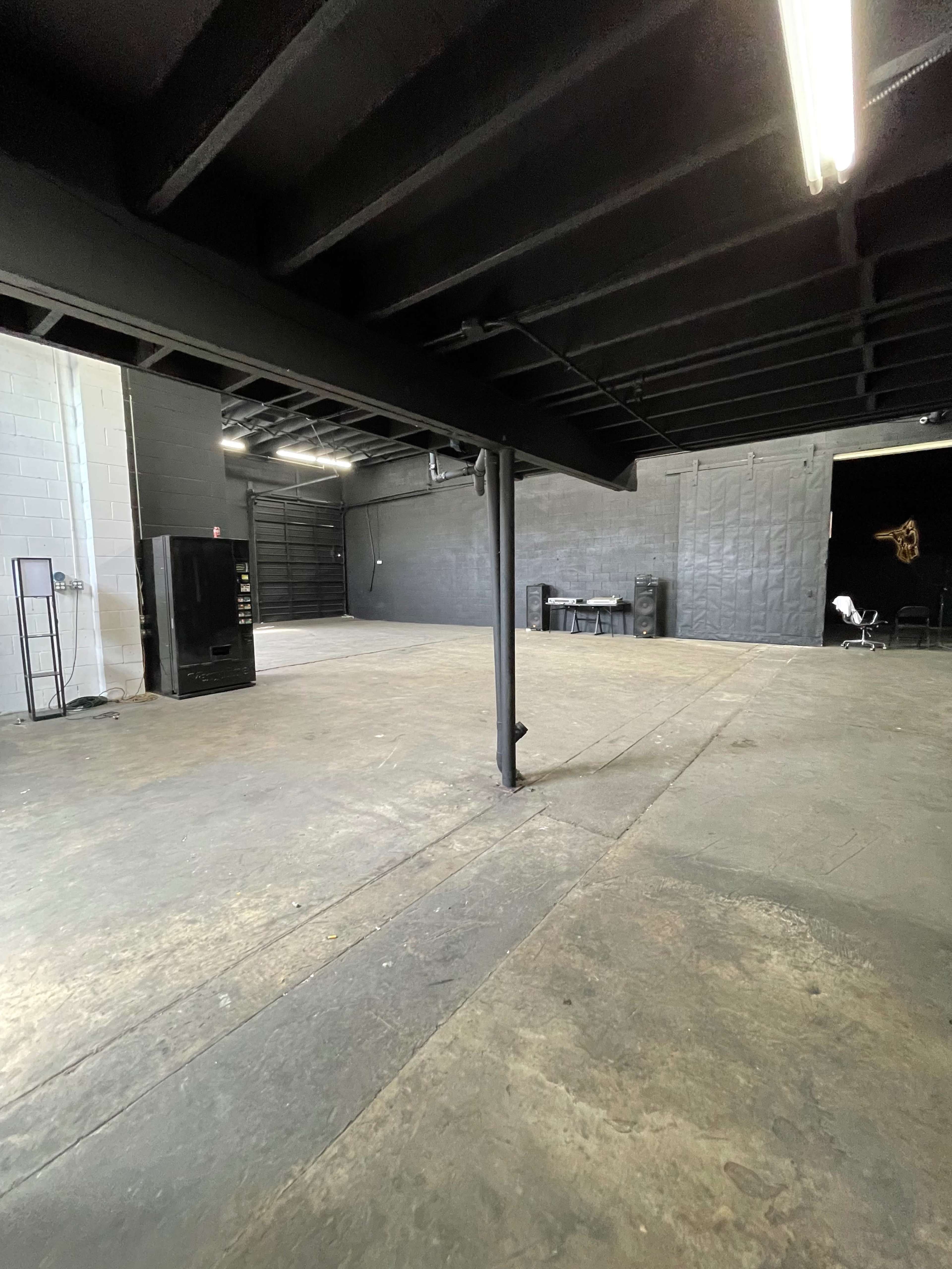 An empty industrial space with a concrete floor and black-painted walls, featuring a refrigerator and a table in the corner.