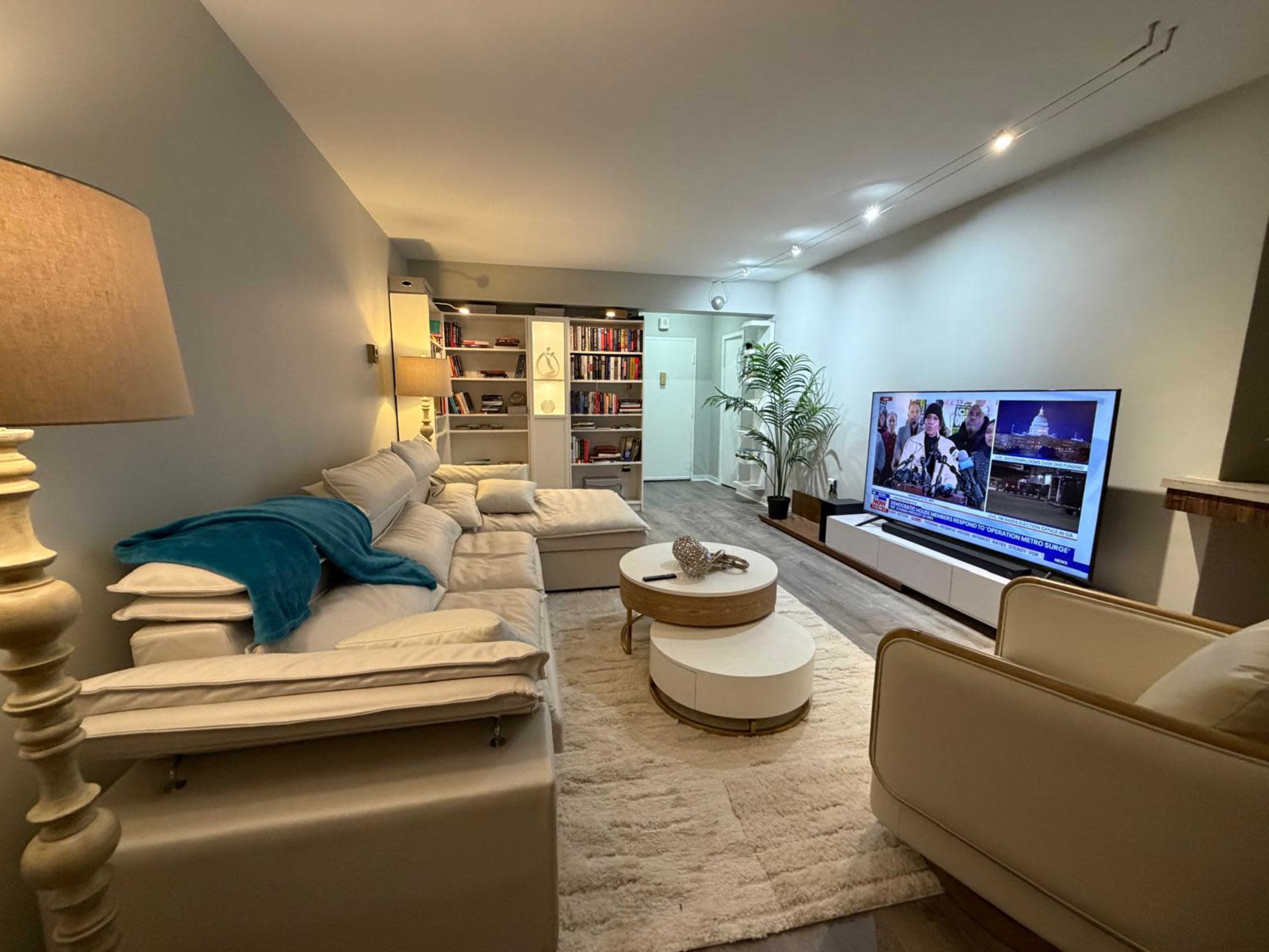 The image shows a modern living room featuring a comfortable seating area, a large television, a bookshelf, and decorative elements like a lamp and a coffee table.