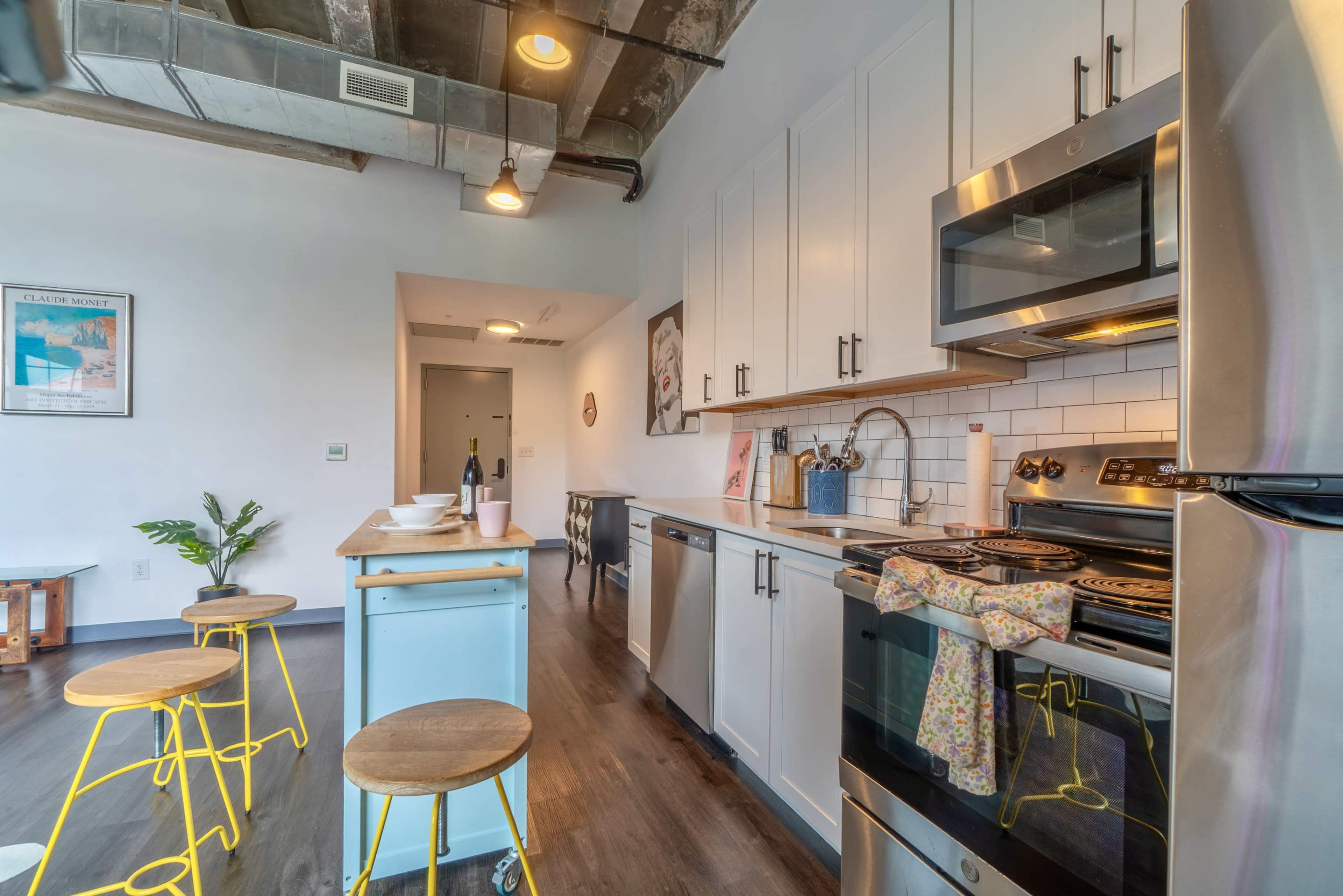 The image shows a modern kitchen with stainless steel appliances, white cabinets, and a small dining area featuring wooden stools and a blue kitchen island.