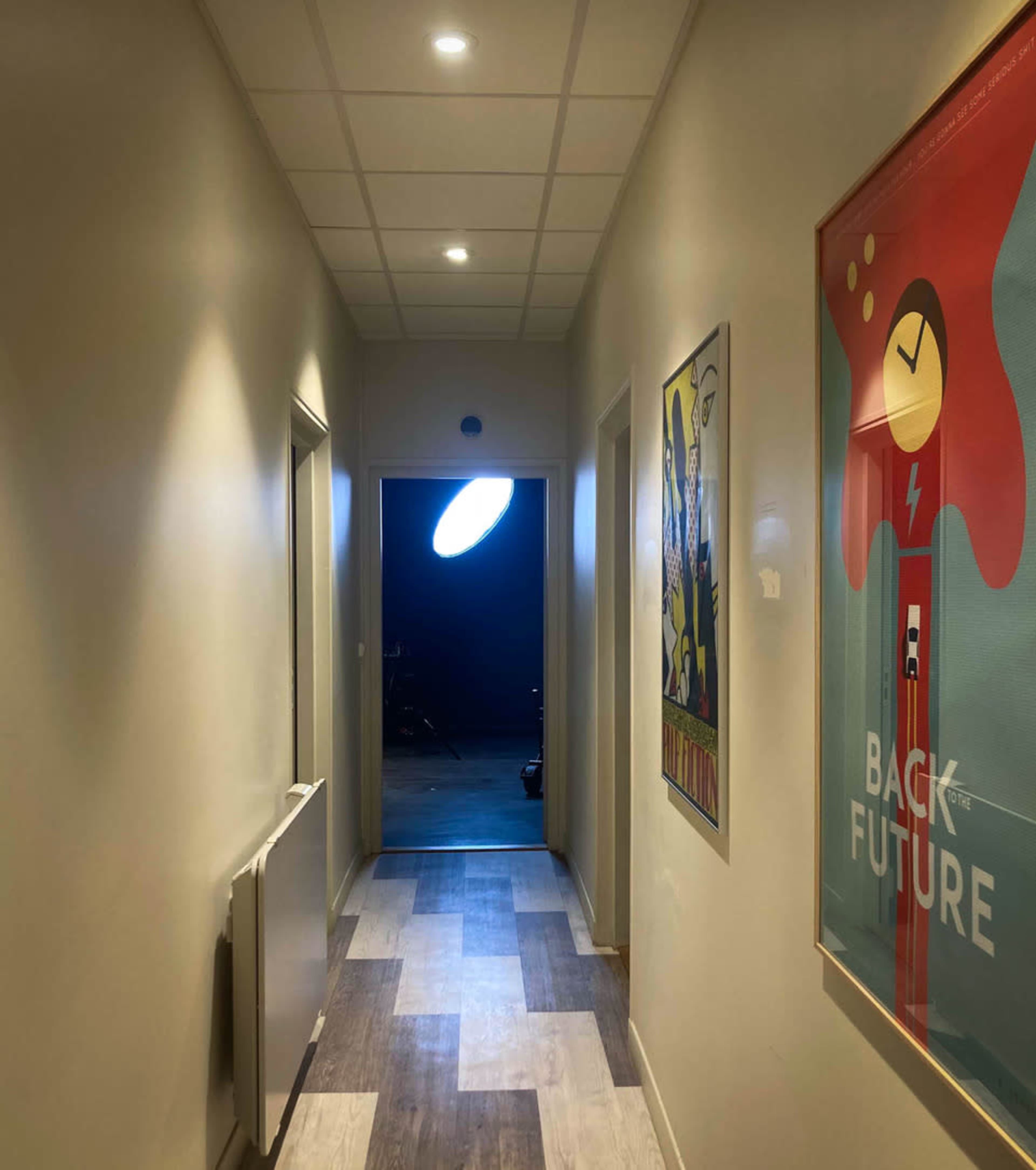 A narrow hallway with two doors on the sides, bright ceiling lights, and a visible opening at the end leading to a darker space.
