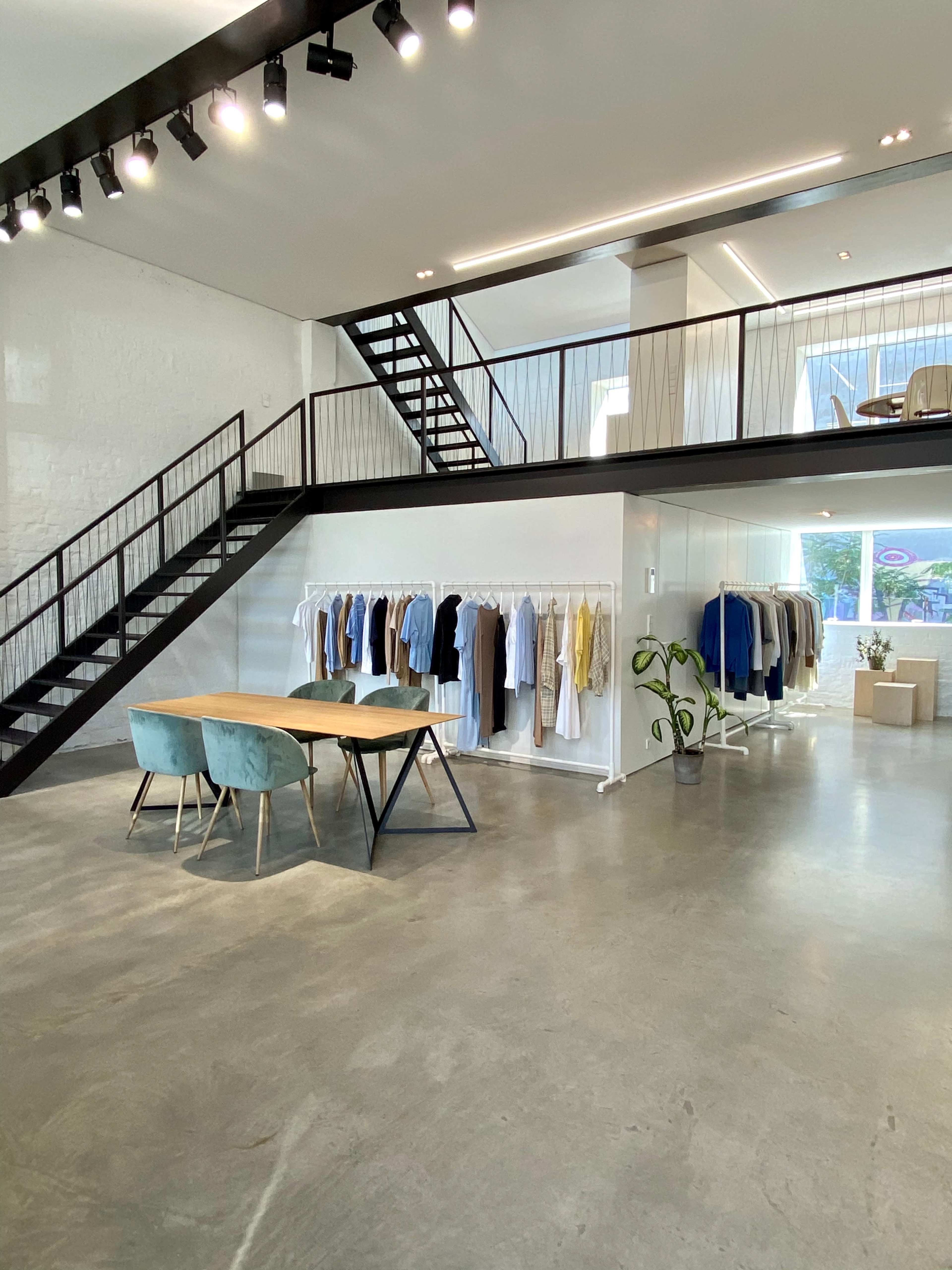 A modern clothing store features a wooden table with blue chairs, hanging garments on racks, and a staircase leading to an upper level.