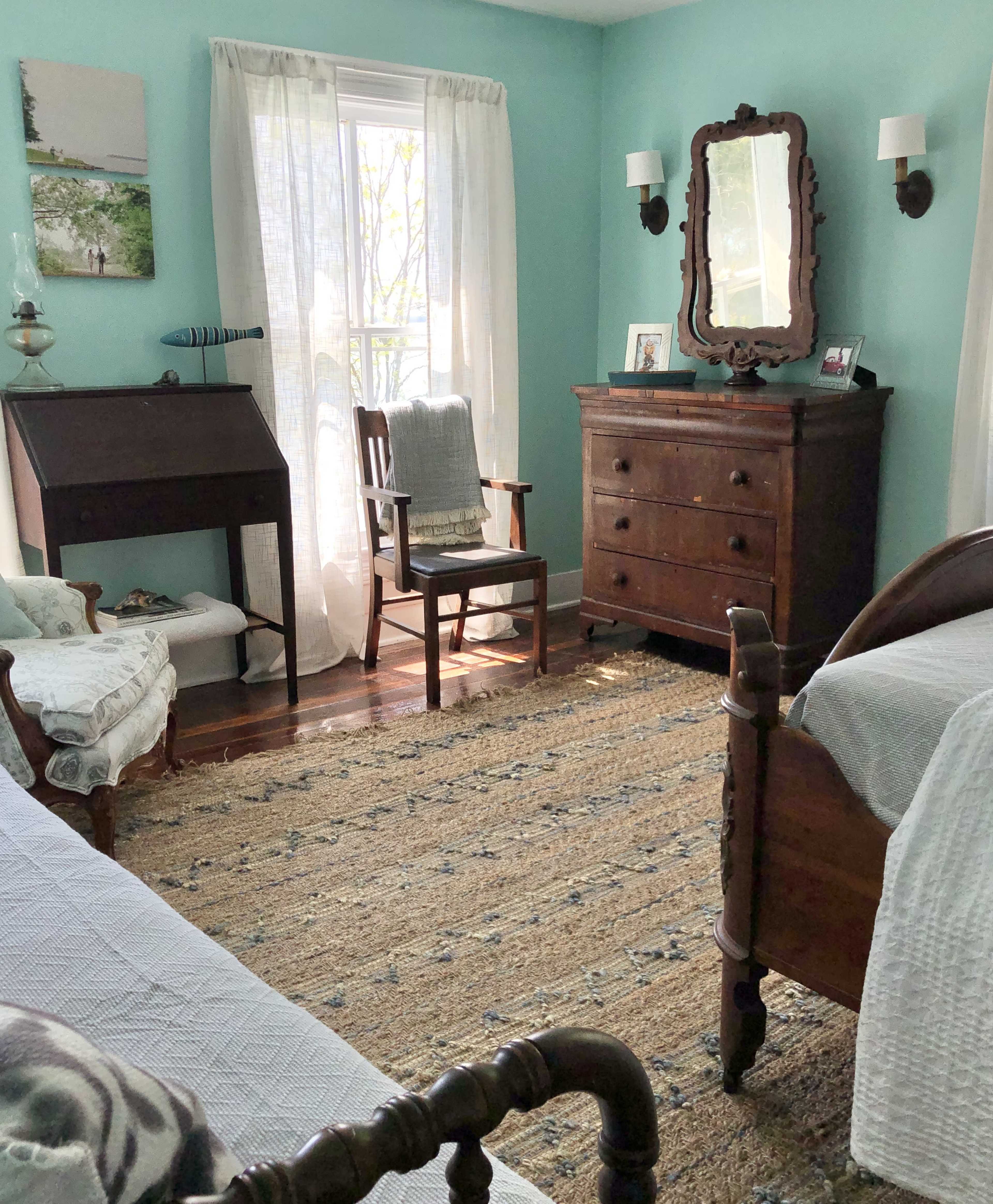 The room features two wooden beds, a dresser, a mirrored vanity, and a large window with sheer curtains, all set against a light blue wall.