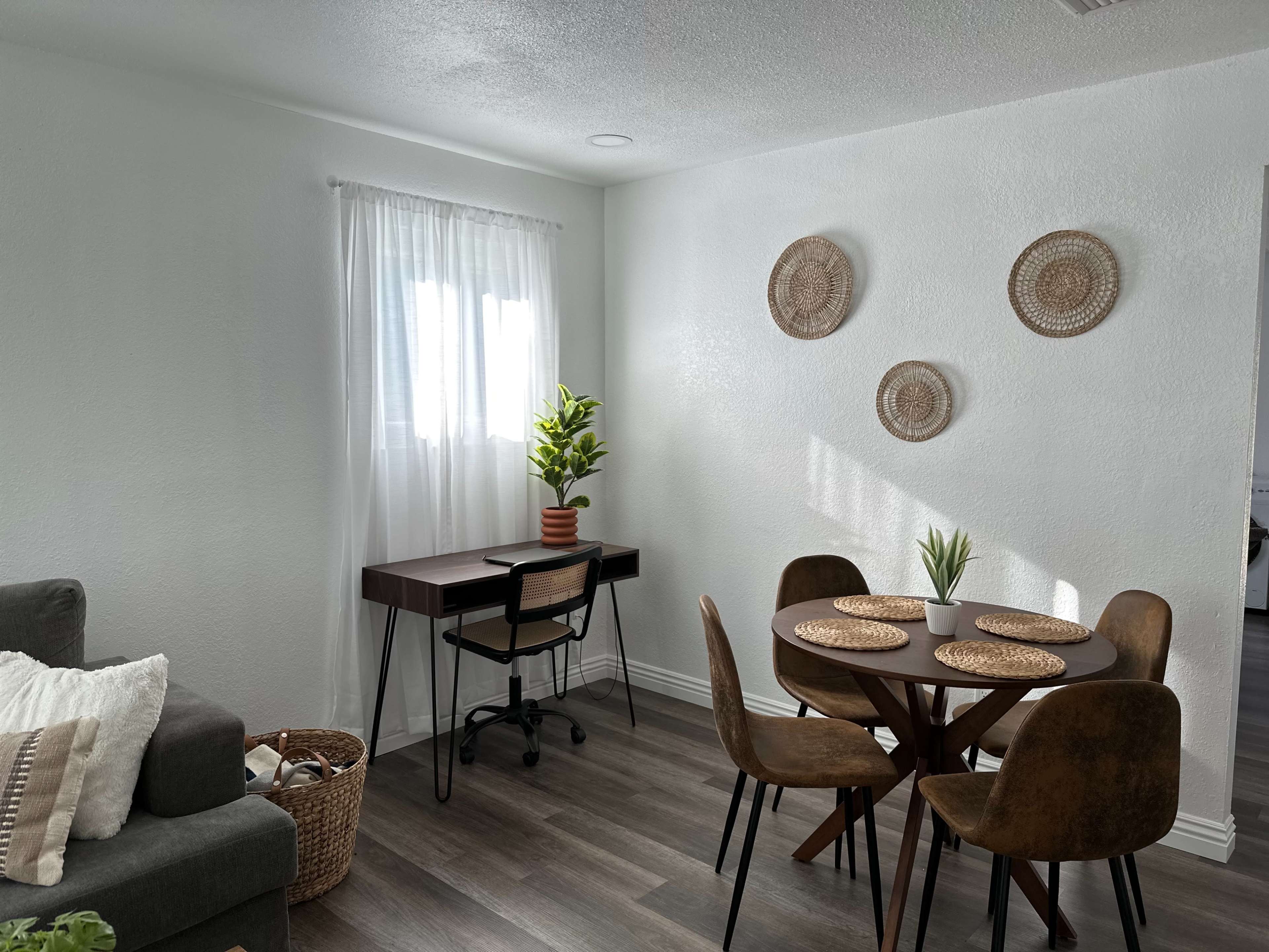 A small room features a gray sofa, a wooden desk with a chair, a round dining table with four brown chairs, and decorative wall hangings.
