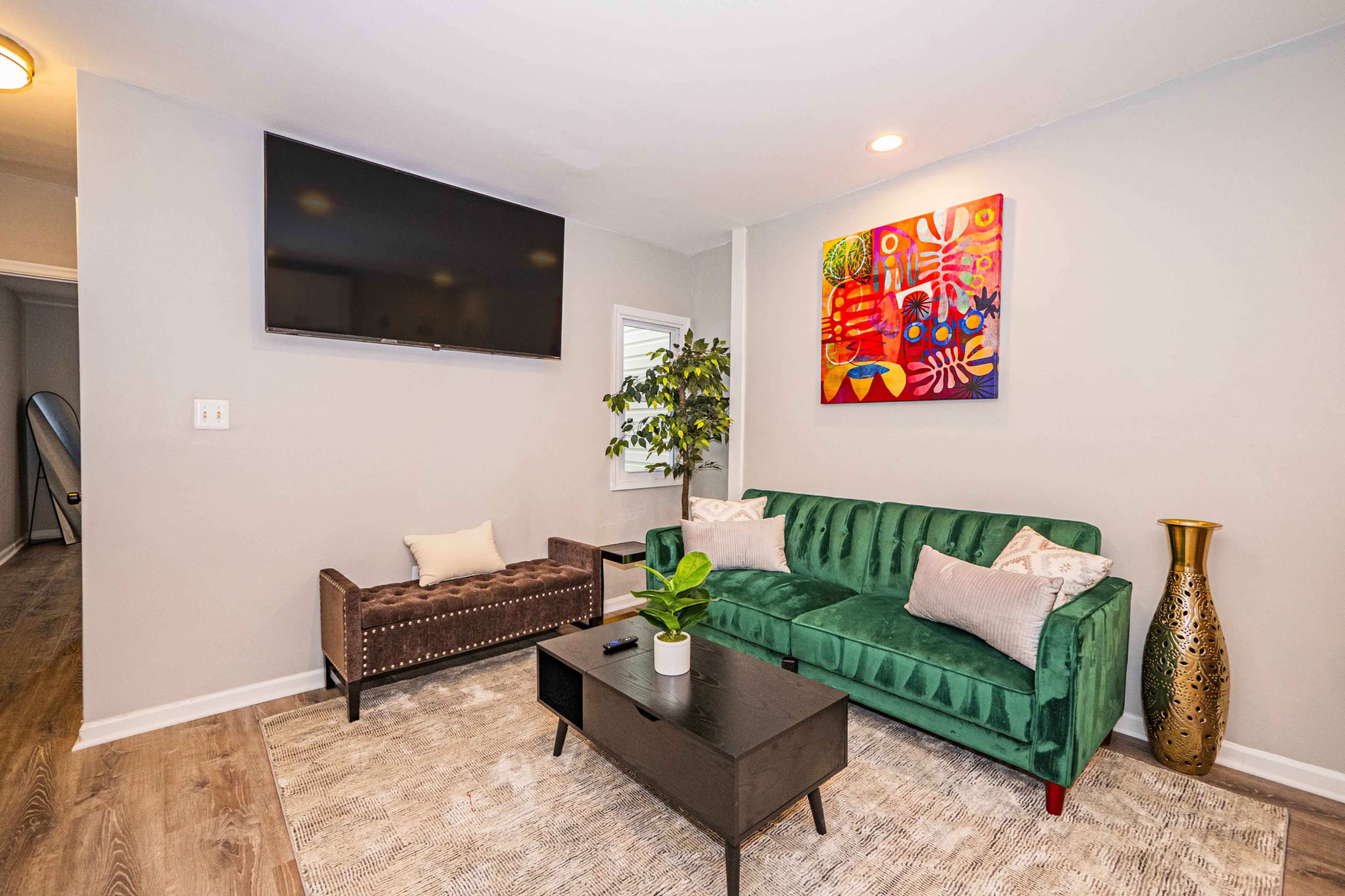 A modern living room features a green velvet sofa, a dark coffee table, a decorative vase, a wall-mounted TV, and a colorful abstract painting.