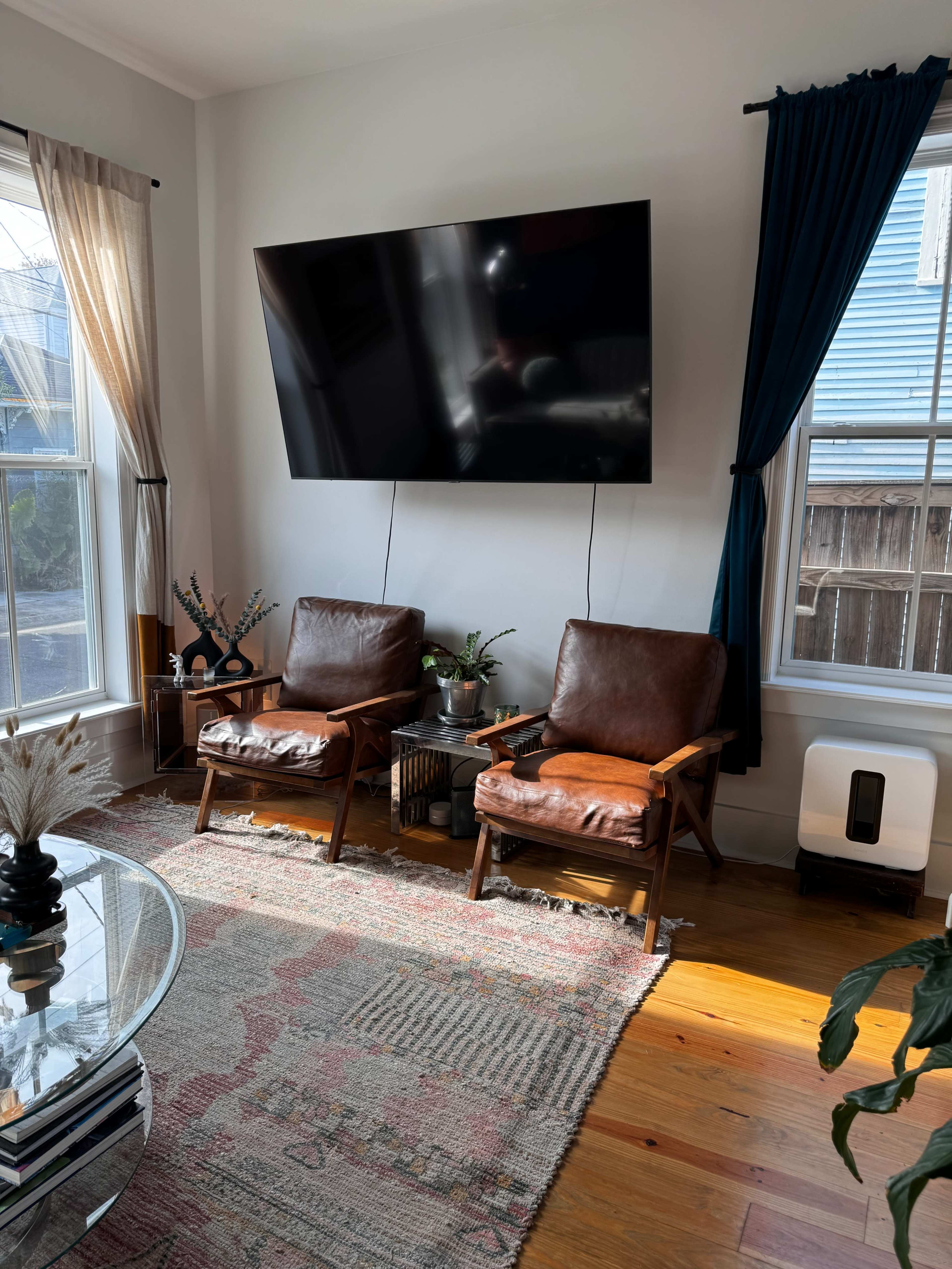 A living room with two brown leather chairs facing a large wall-mounted television, surrounded by windows with light curtains and a patterned rug on the floor.